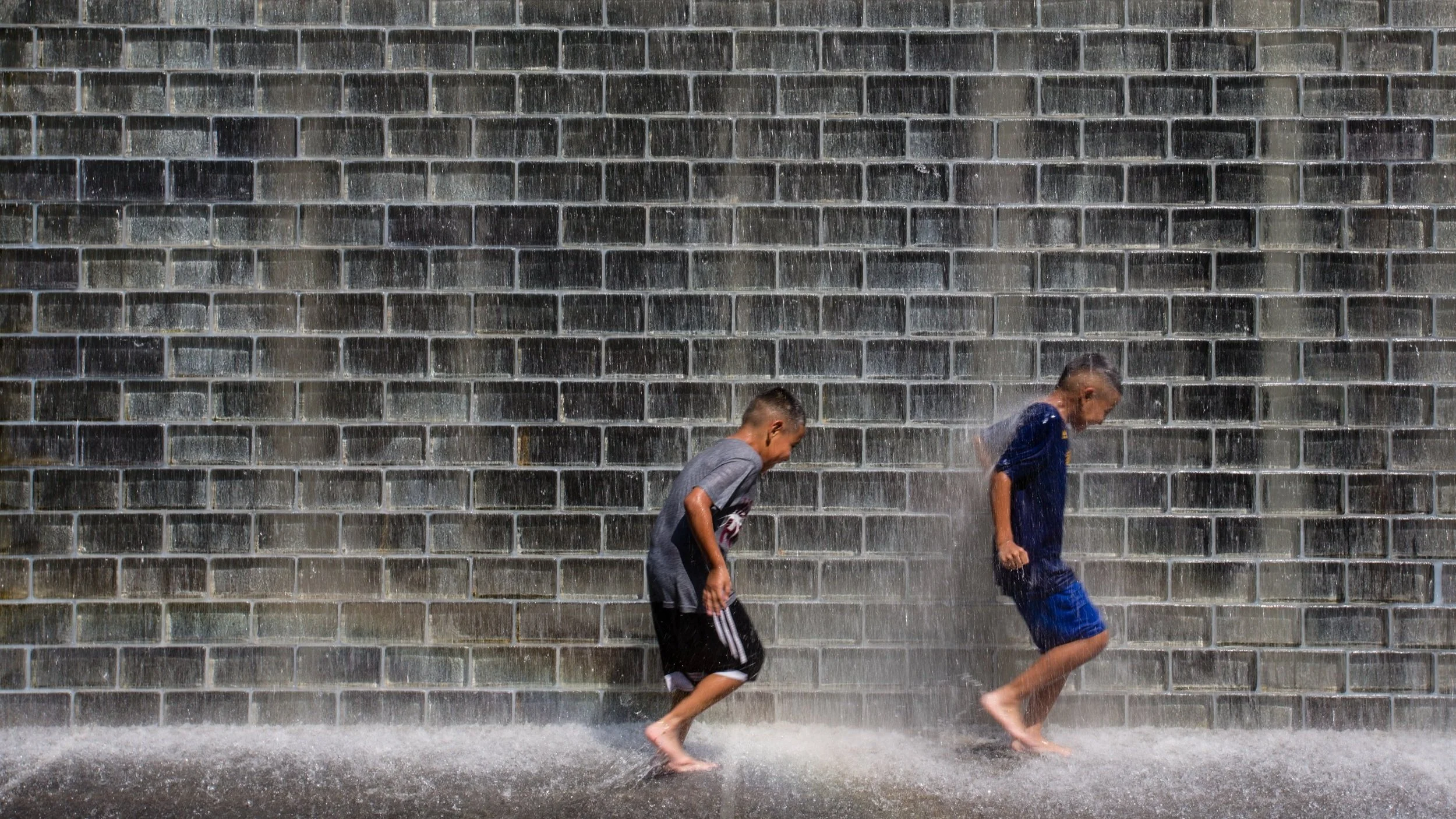 Two boys playing and splashing water near a brick wall.