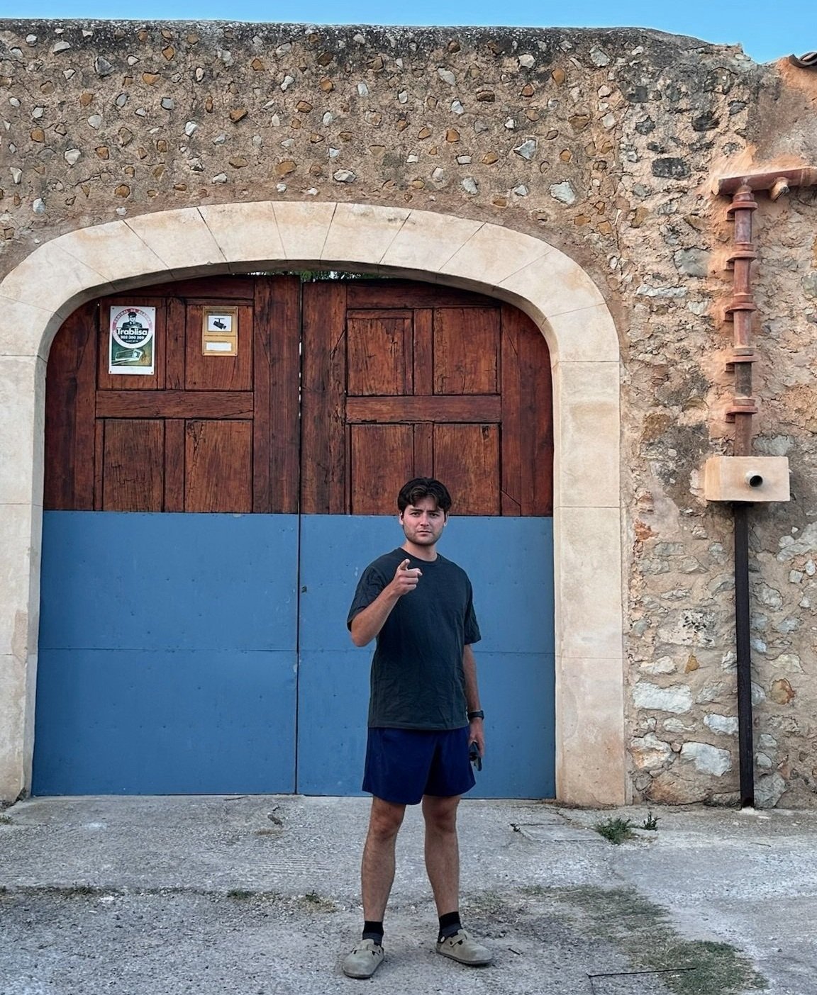 Young man standing in front of a large wooden gate with a stone and stucco wall behind him, pointing at the camera.