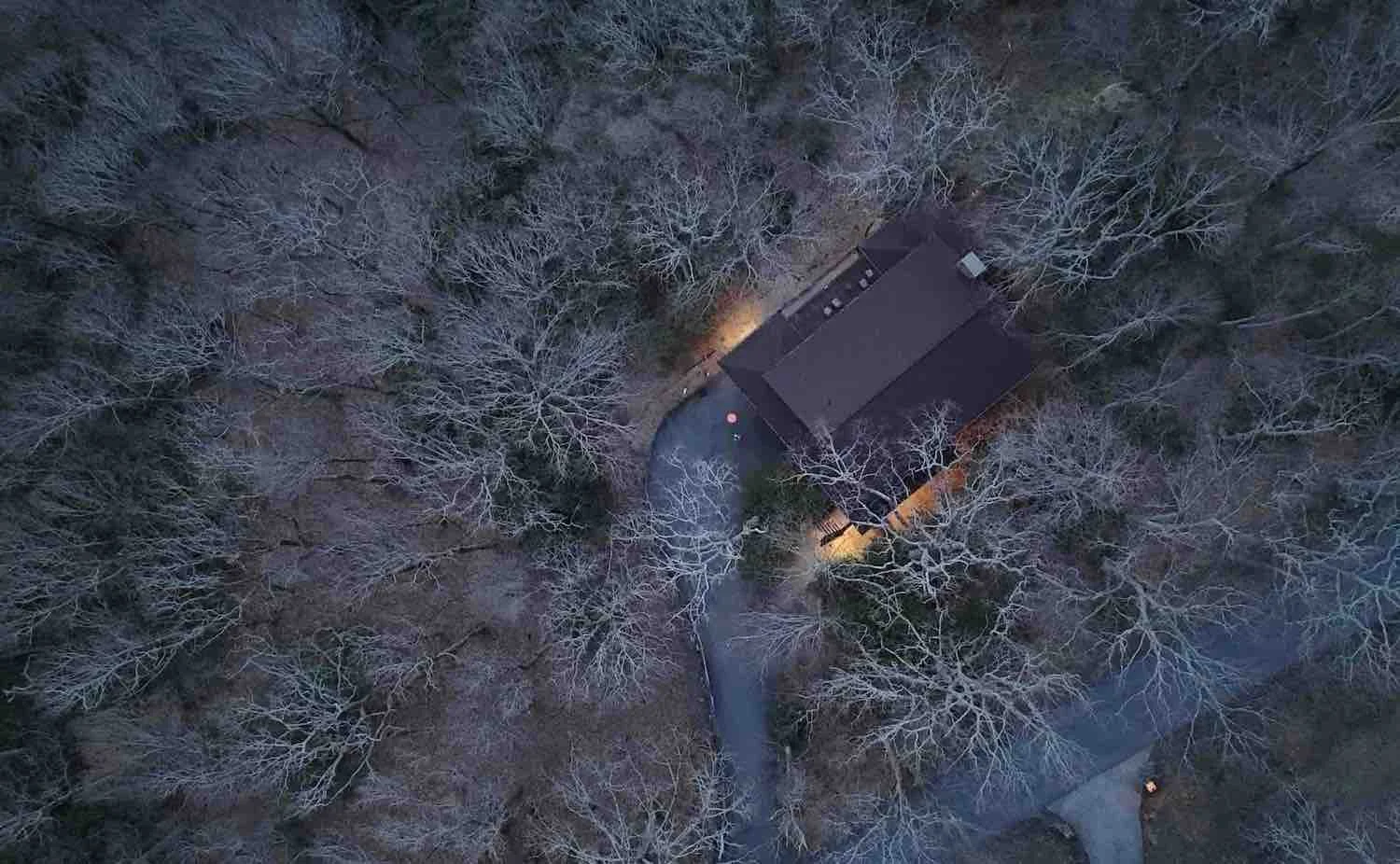 Aerial view of a house surrounded by leafless trees during winter, with a driveway leading to the house and warm outdoor lighting.