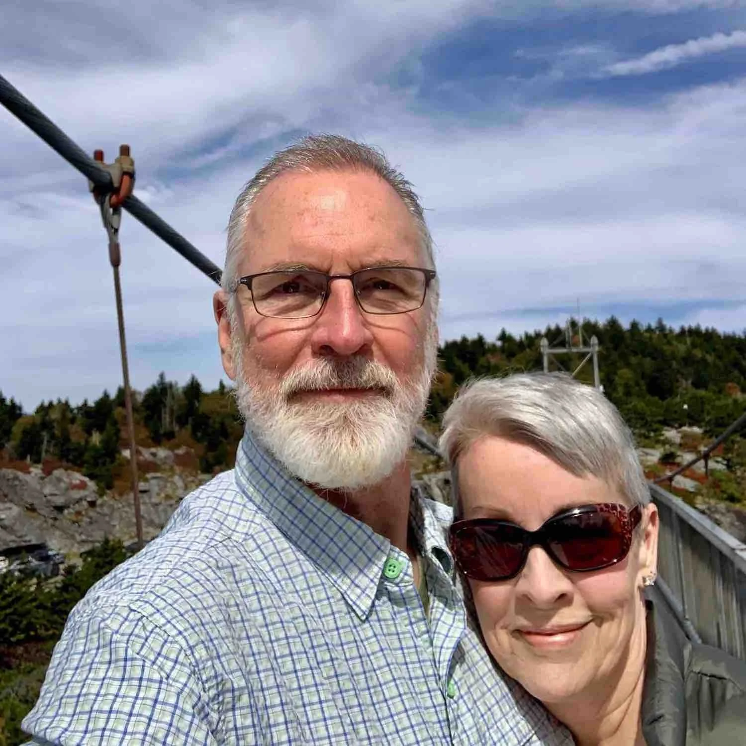 A smiling older man with glasses, a beard, and a checkered shirt, and a woman with short gray hair, sunglasses, and earrings, taking a selfie on a bridge with a rocky hillside and electrical towers in the background.