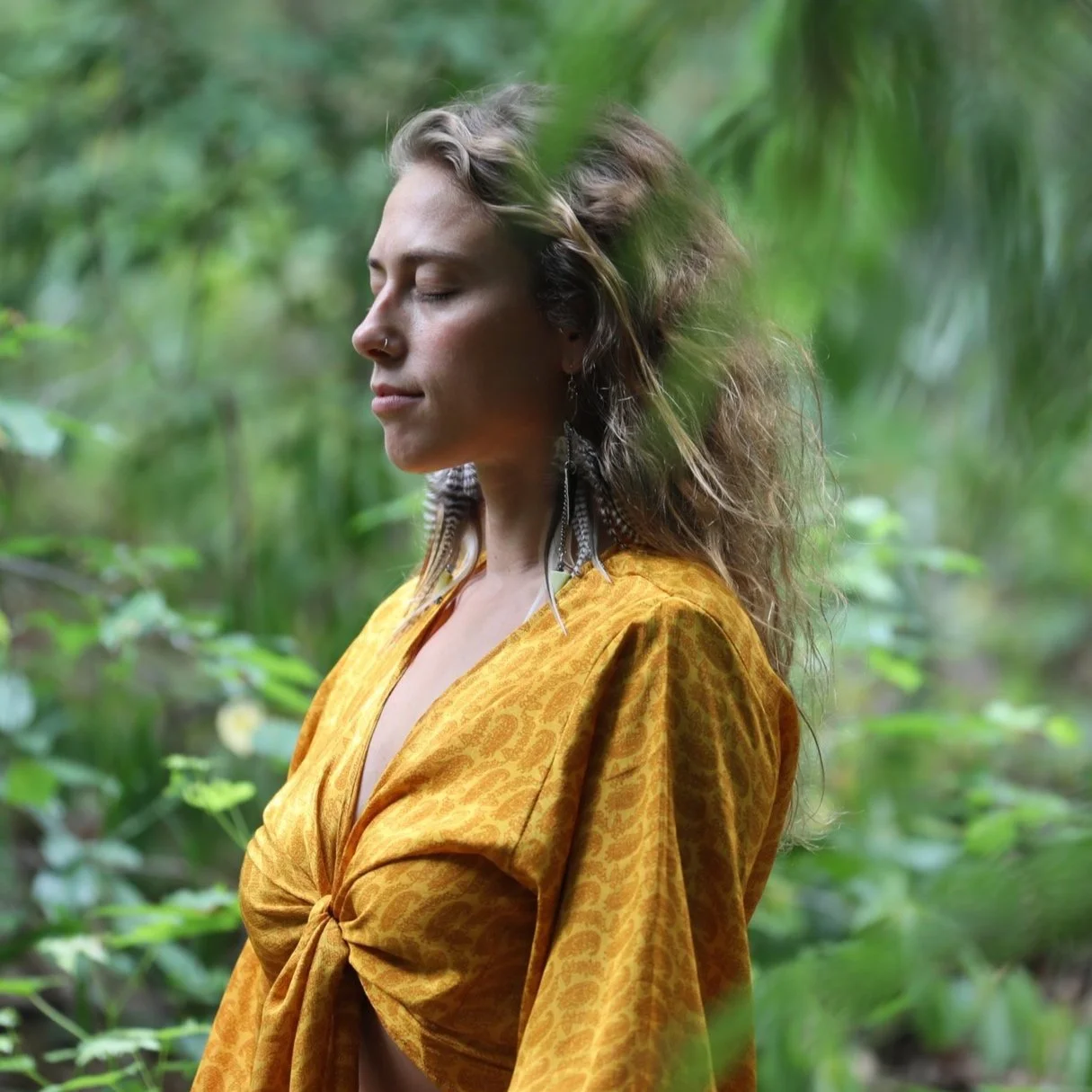 A woman with closed eyes and long earrings stands in a green forest, wearing a mustard-colored, patterned blouse with a knot in the front.