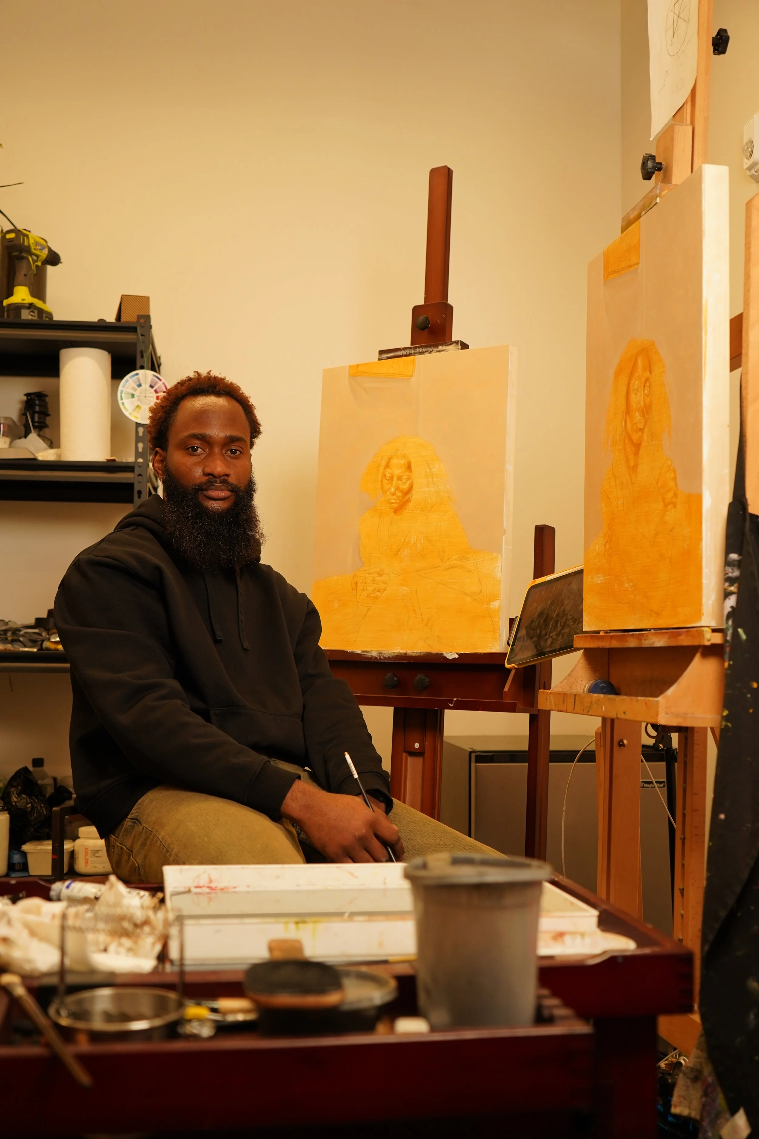 A man with a beard sitting in an art studio, with two unfinished paintings of a woman with long hair on easels behind him.