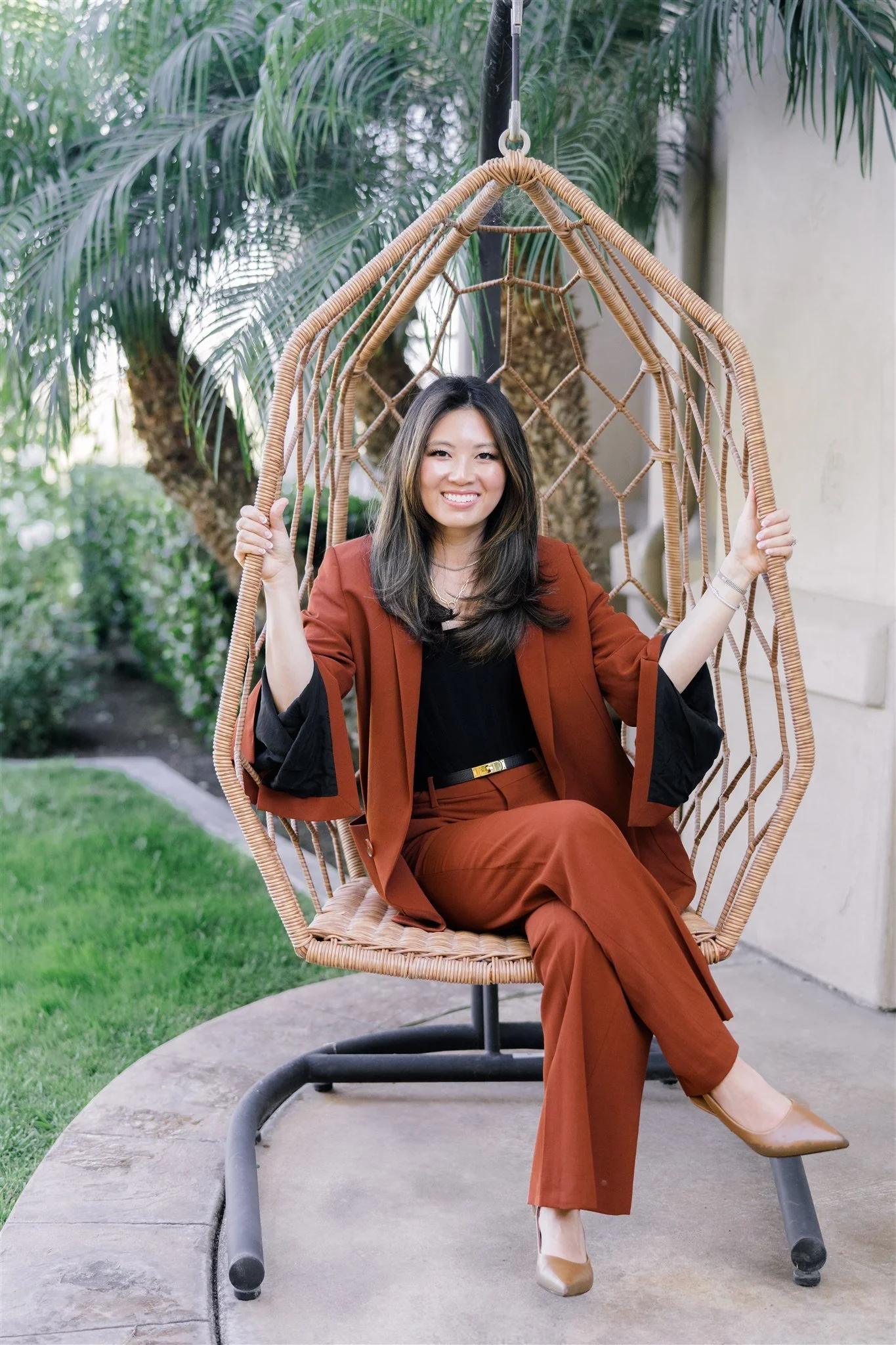 A woman in a rust-colored suit sitting on a hanging wicker chair outdoors, smiling, with palm trees and greenery in the background.