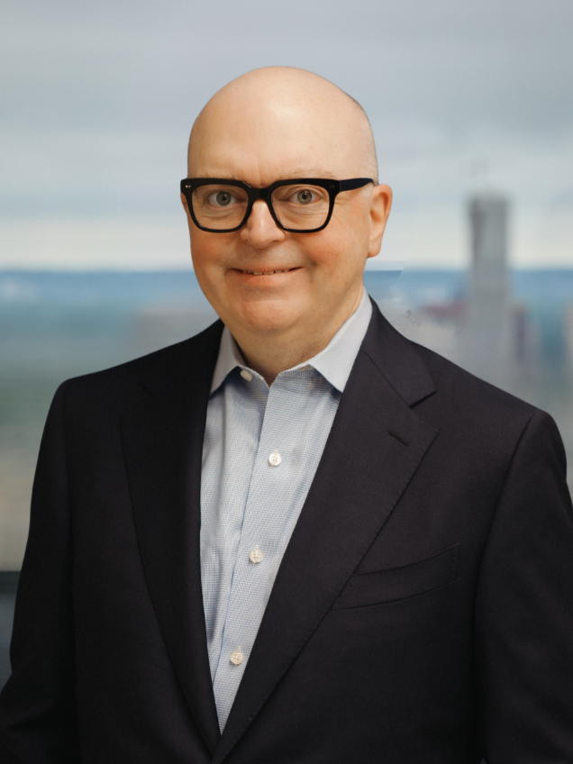 Black-and-white portrait of a bald man with glasses, wearing a suit and a button-up shirt, smiling at the camera against a dark textured background.