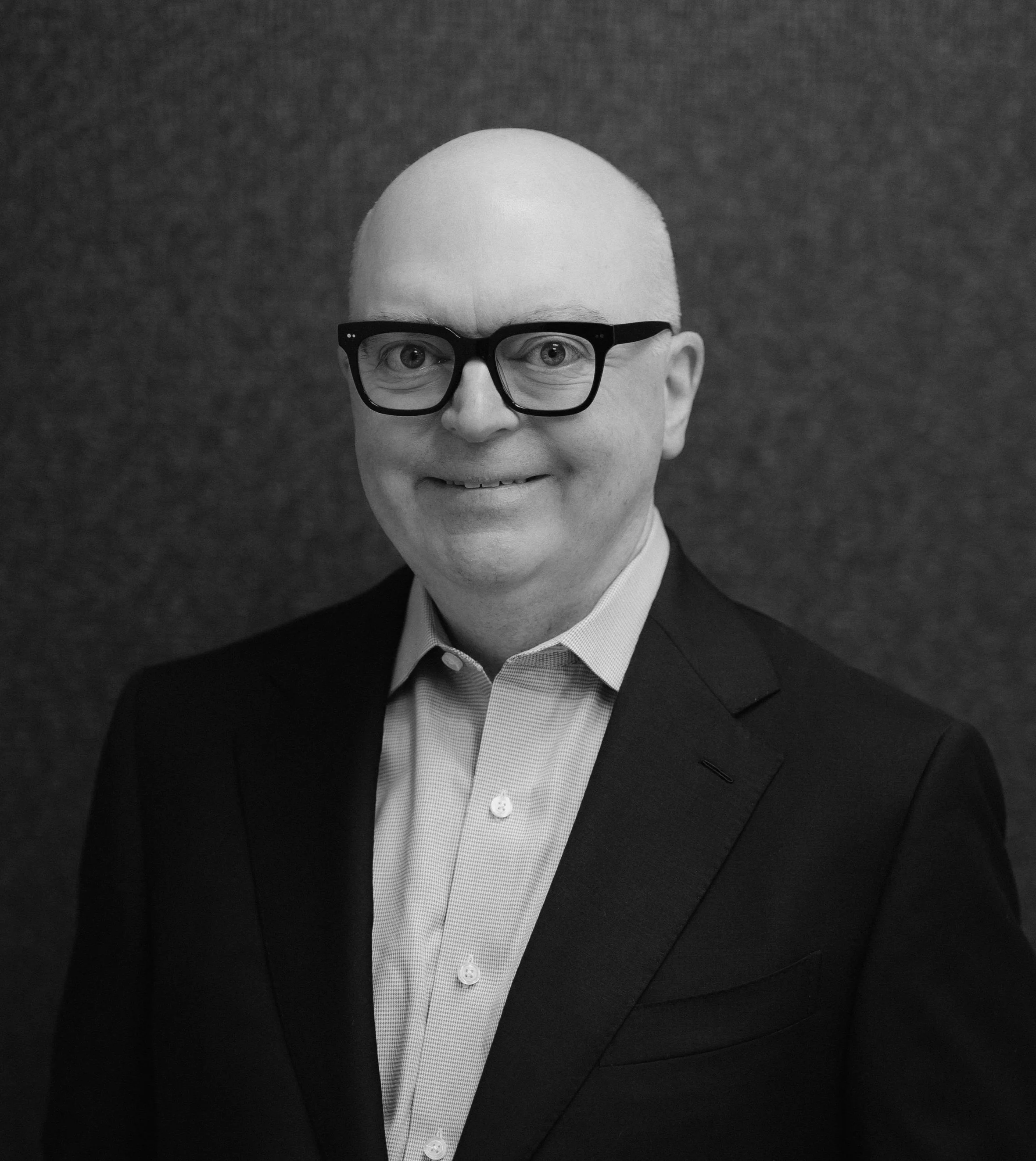 Black-and-white portrait of a bald man with glasses, wearing a suit and a button-up shirt, smiling at the camera against a dark textured background.