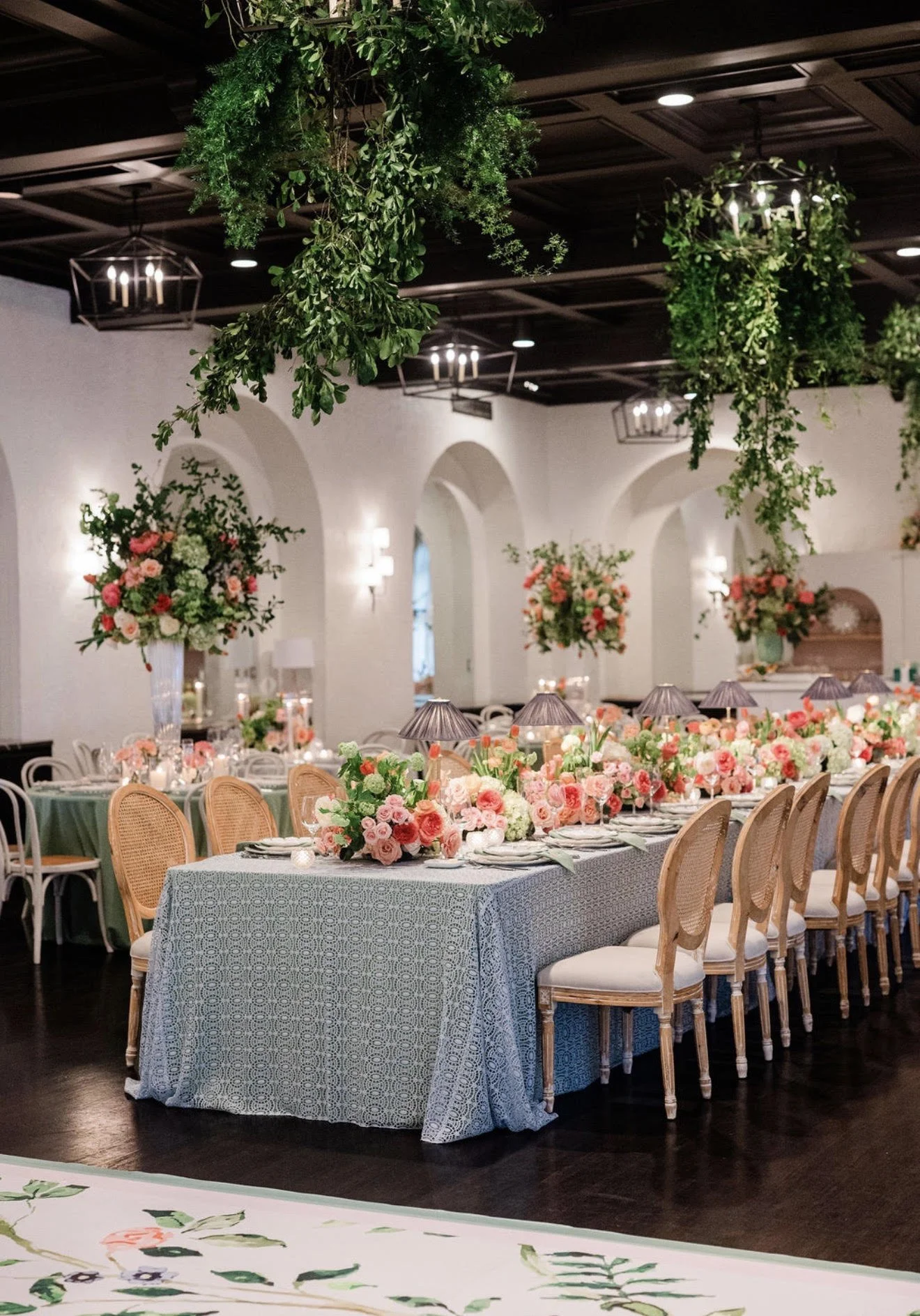 An elegantly decorated indoor event space with long table set for a formal gathering, adorned with floral centerpieces, garlands with flowers, and greenery hanging from the ceiling. The room features high ceilings, arches, wall-mounted lighting, and 