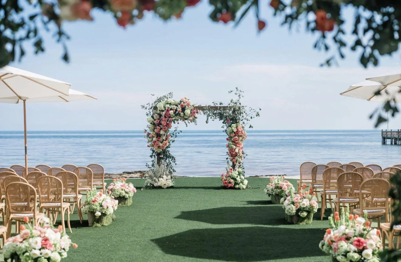 Beachside wedding ceremony setup with floral arch, chairs, and umbrellas overlooking the ocean.