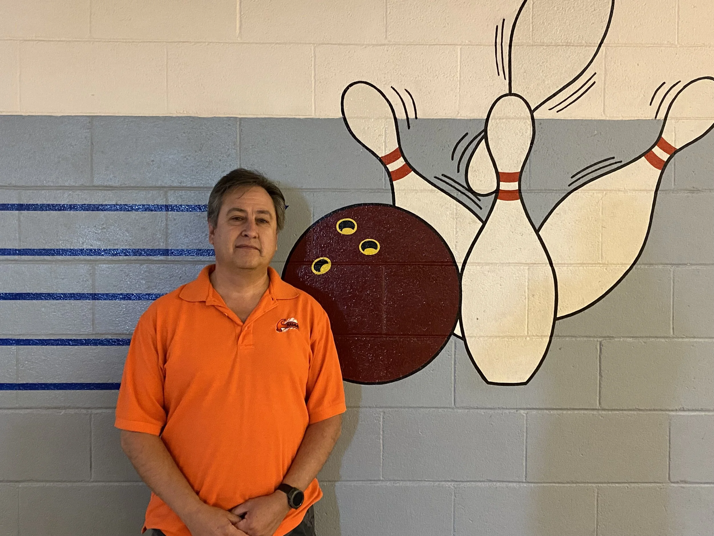 Mark Archuleta, in an orange polo shirt, standing in front of a wall w/ a bowling ball knocking down 4 pins.
