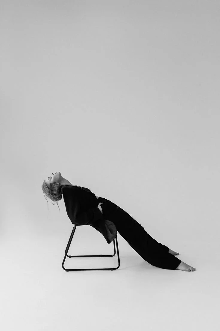 Black and white photo of a person sitting backward on a chair, leaning back with their head tilted upward and smiling, legs extended out on the floor, against a plain background.