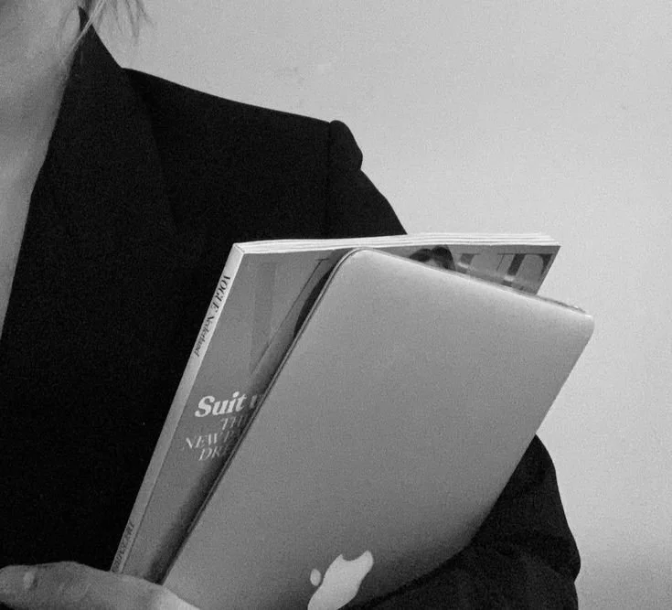 Person in a dark blazer holding a closed MacBook laptop with an open magazine on top in black and white.