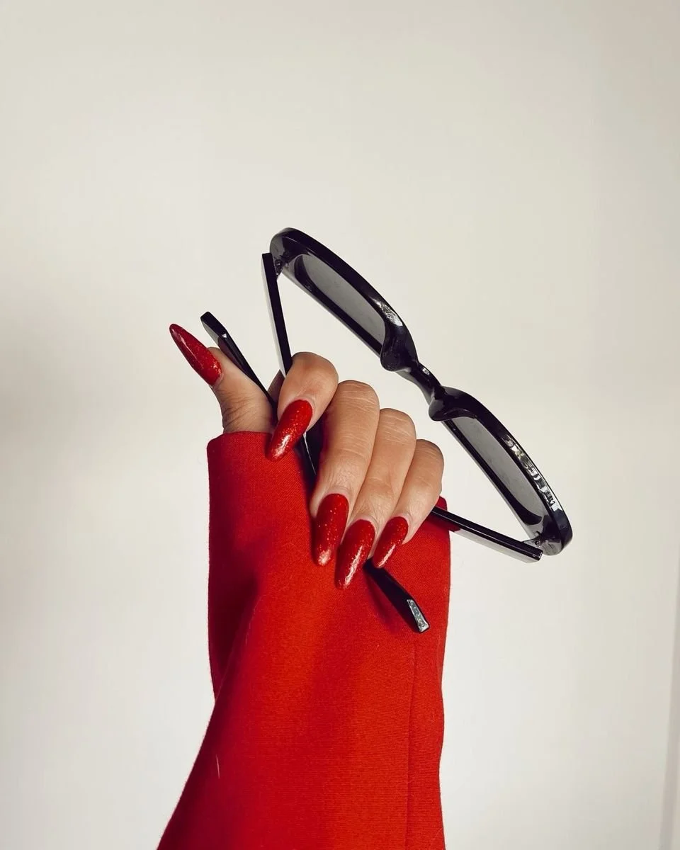 Hand with red nail polish and red sleeve holding black sunglasses against a plain white background.