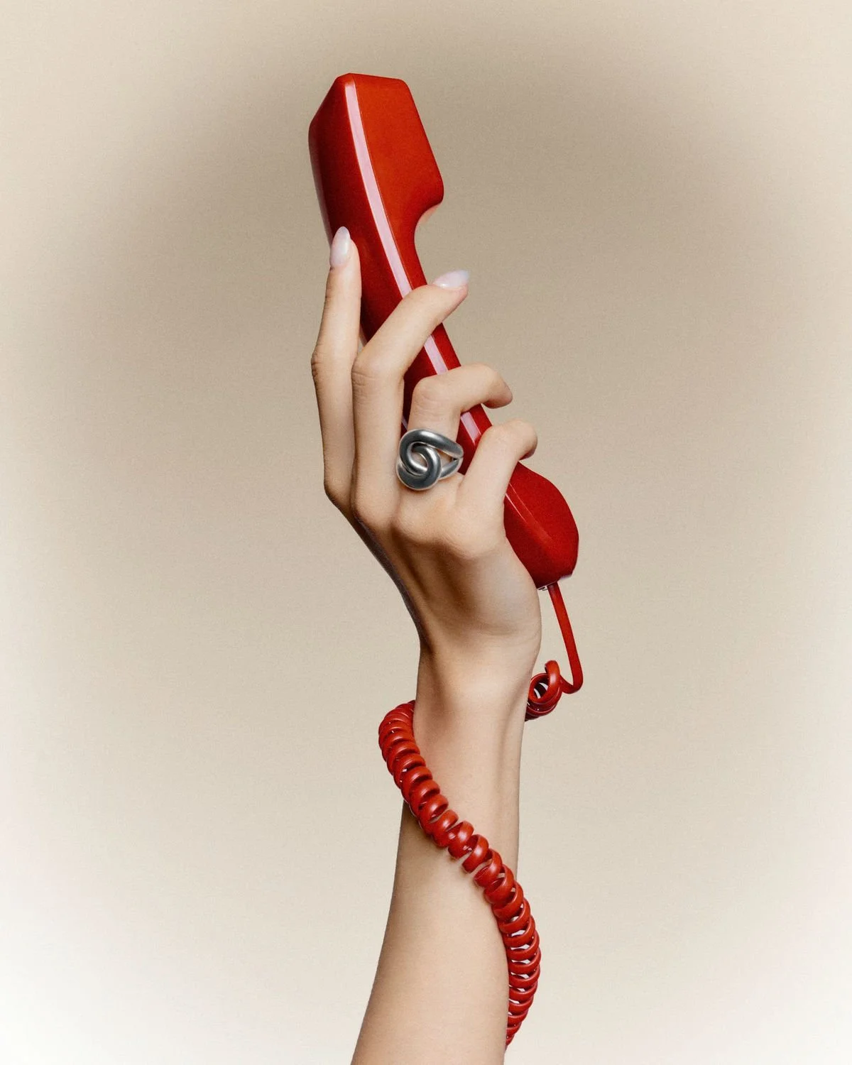 A hand holding a red vintage rotary telephone receiver, with a coiled red cord, and wearing a silver spiral ring.
