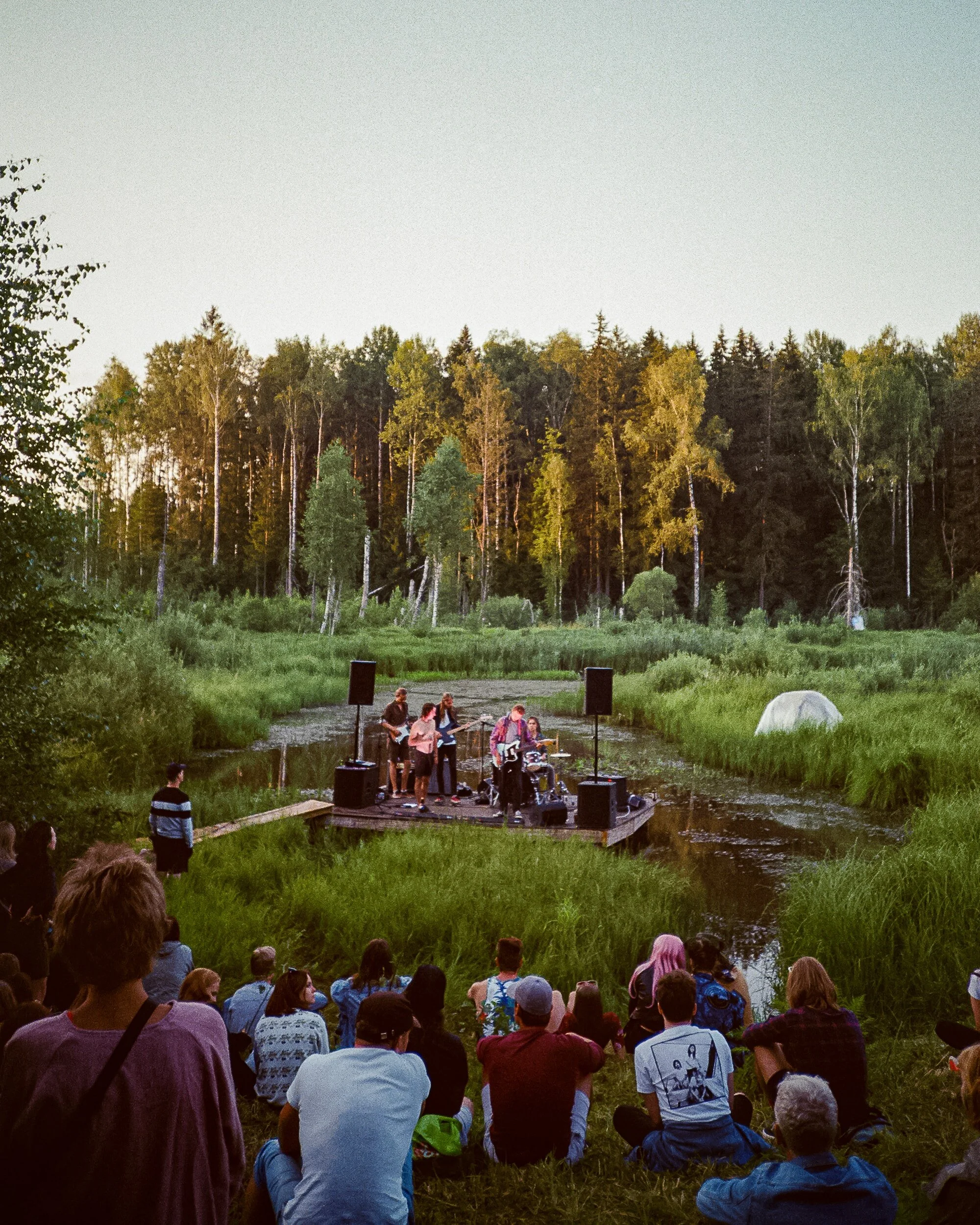 Group of young adults sitting on hill overlooking a lake and a band playing on a stage