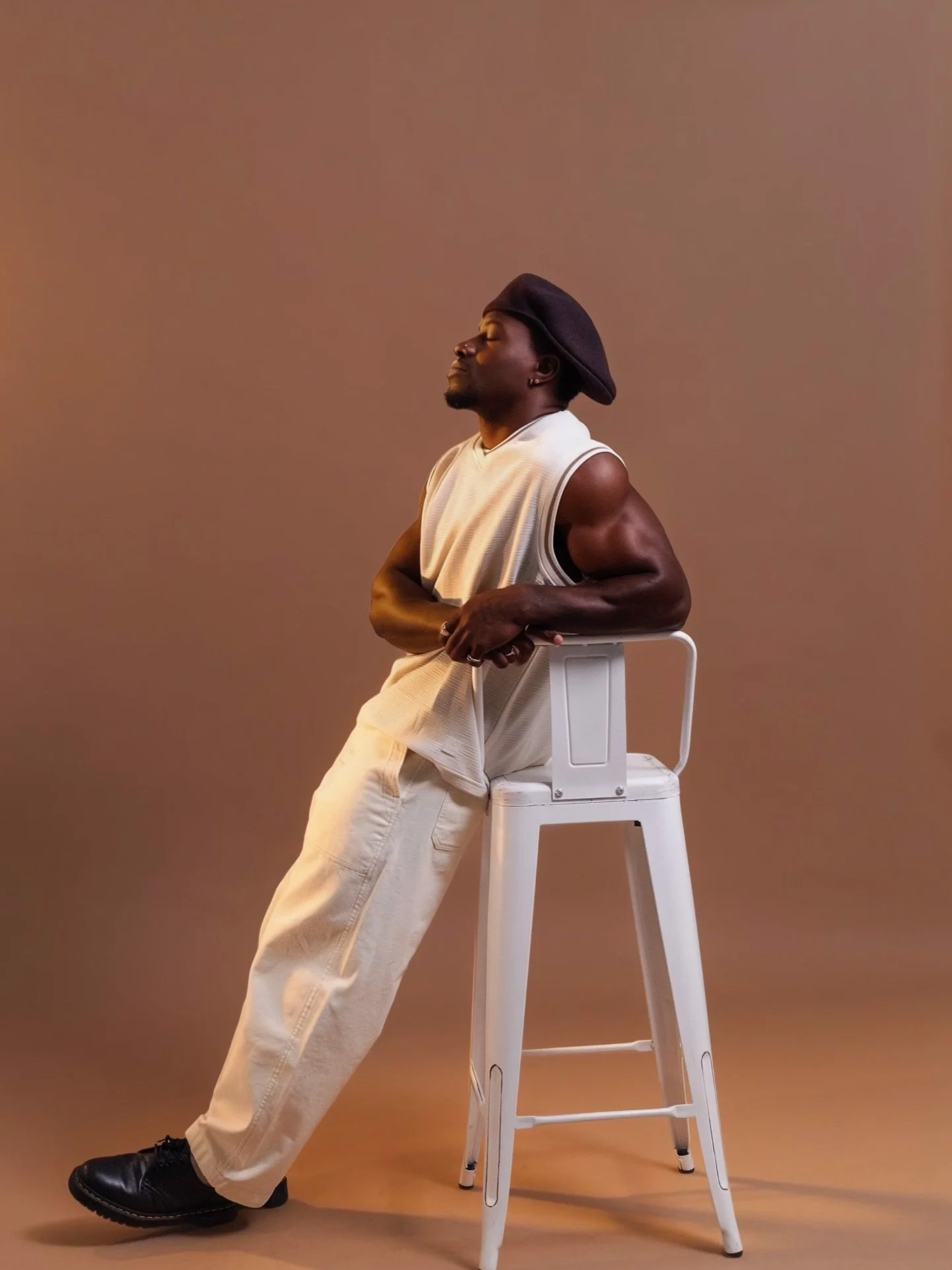 Model in a white tank top and cream trousers leaning on a white stool against a warm brown studio backdrop, shot at Photography Club