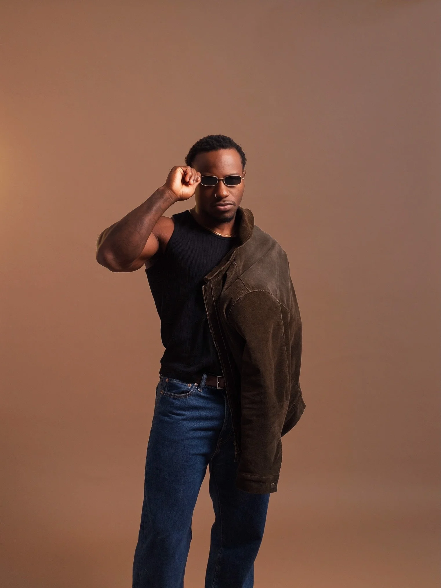 Model in a black T-shirt and jeans with a dark jacket over his shoulder, adjusting his sunglasses against a warm brown studio backdrop, shot at Photography Club
