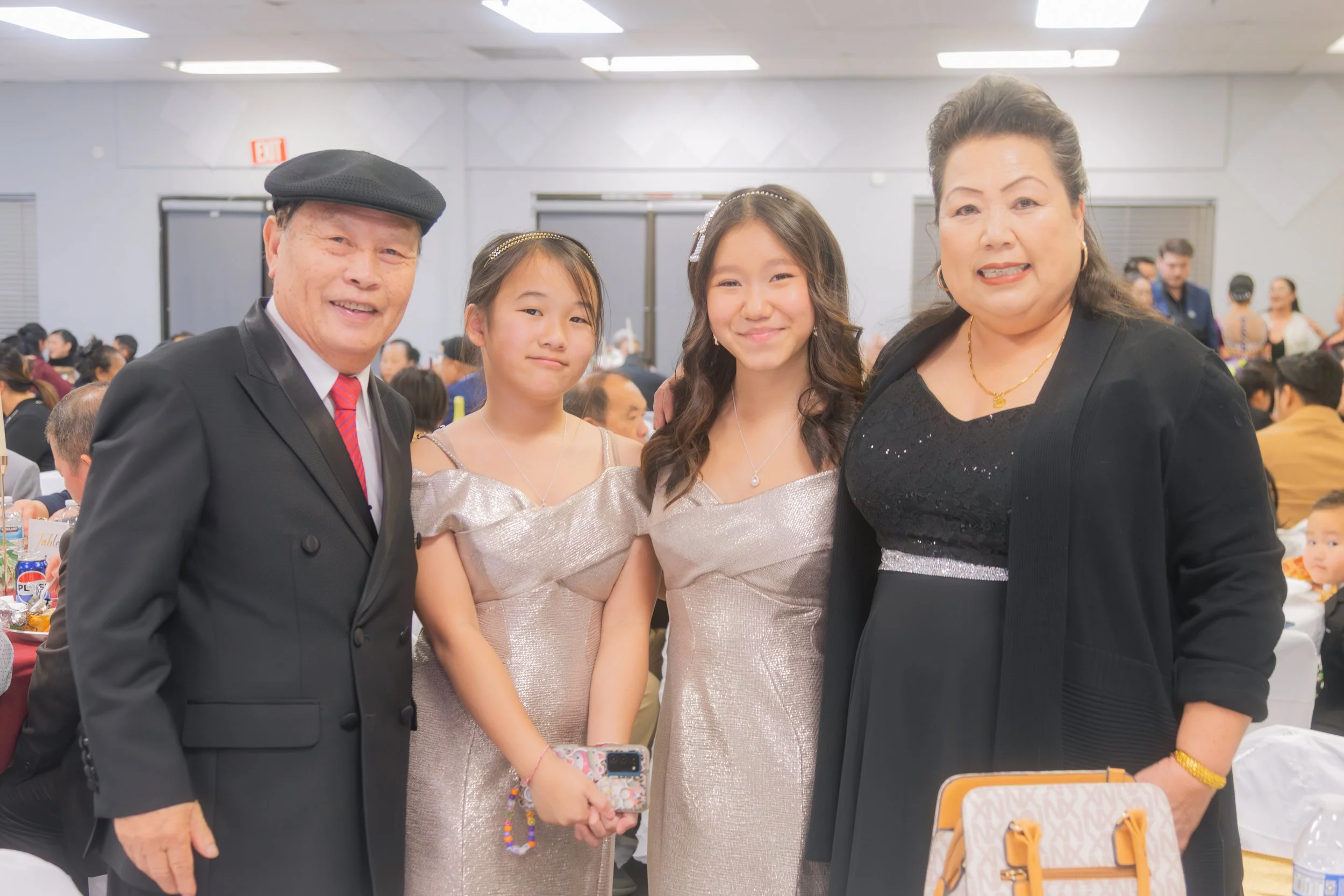 A family of four smiling at a formal event, with two young girls in matching dresses, a man in a black suit and red tie, and a woman in a black dress with jewelry, in a crowded banquet hall.