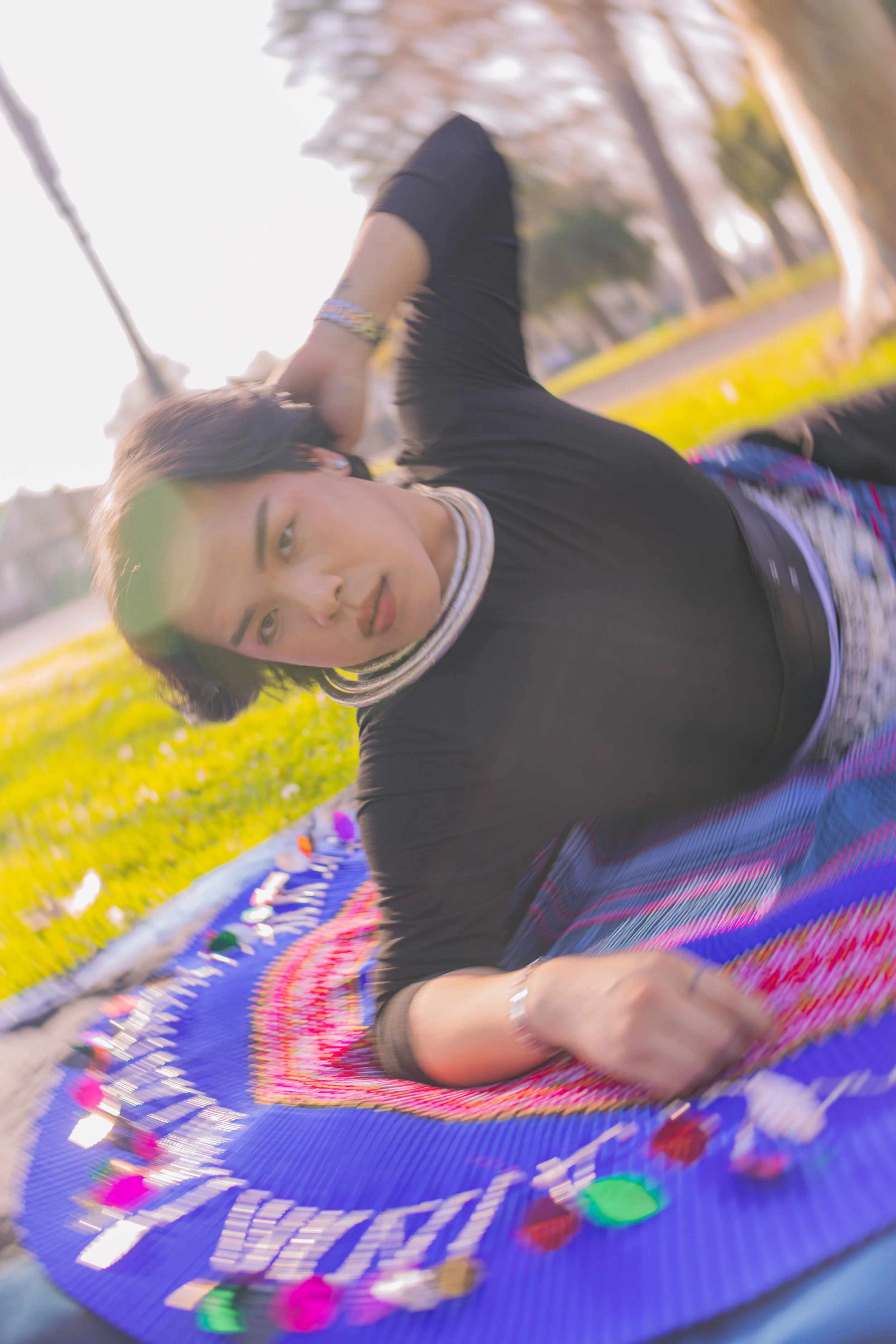 A woman with dark hair and a striped turtleneck shirt lying on colorful layered blanket outdoors on green grass, with trees and a bright sky in the background.