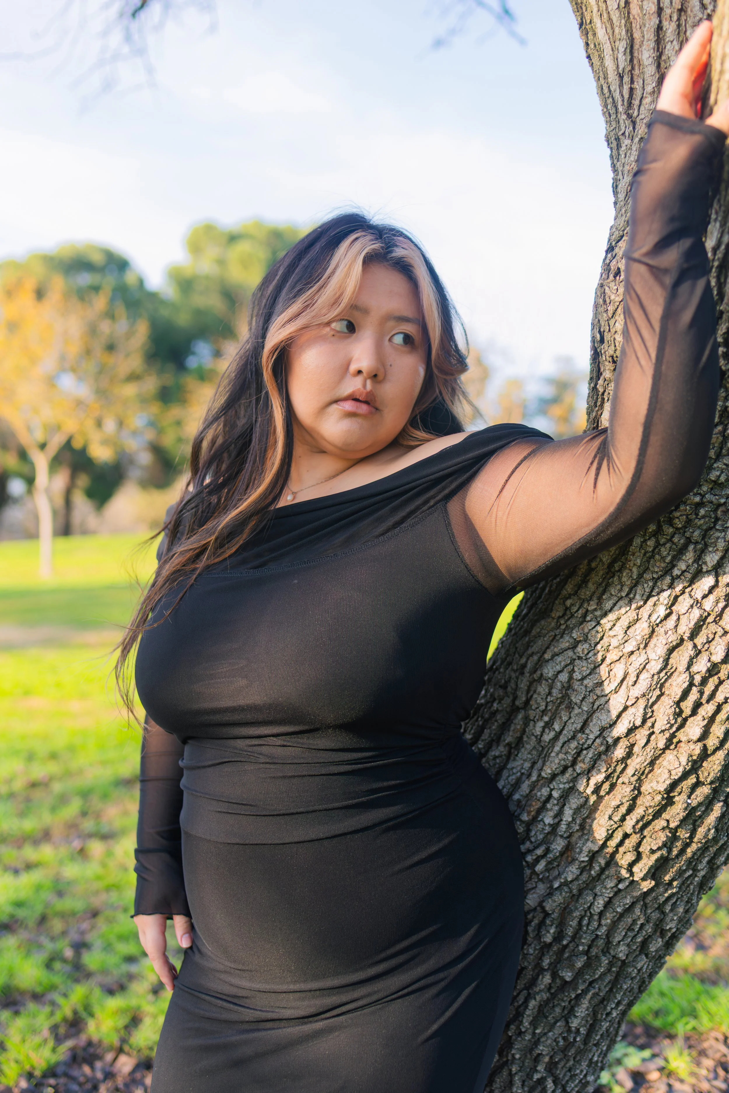 A woman with long wavy hair and a black dress leans against a tree in a park on a sunny day.