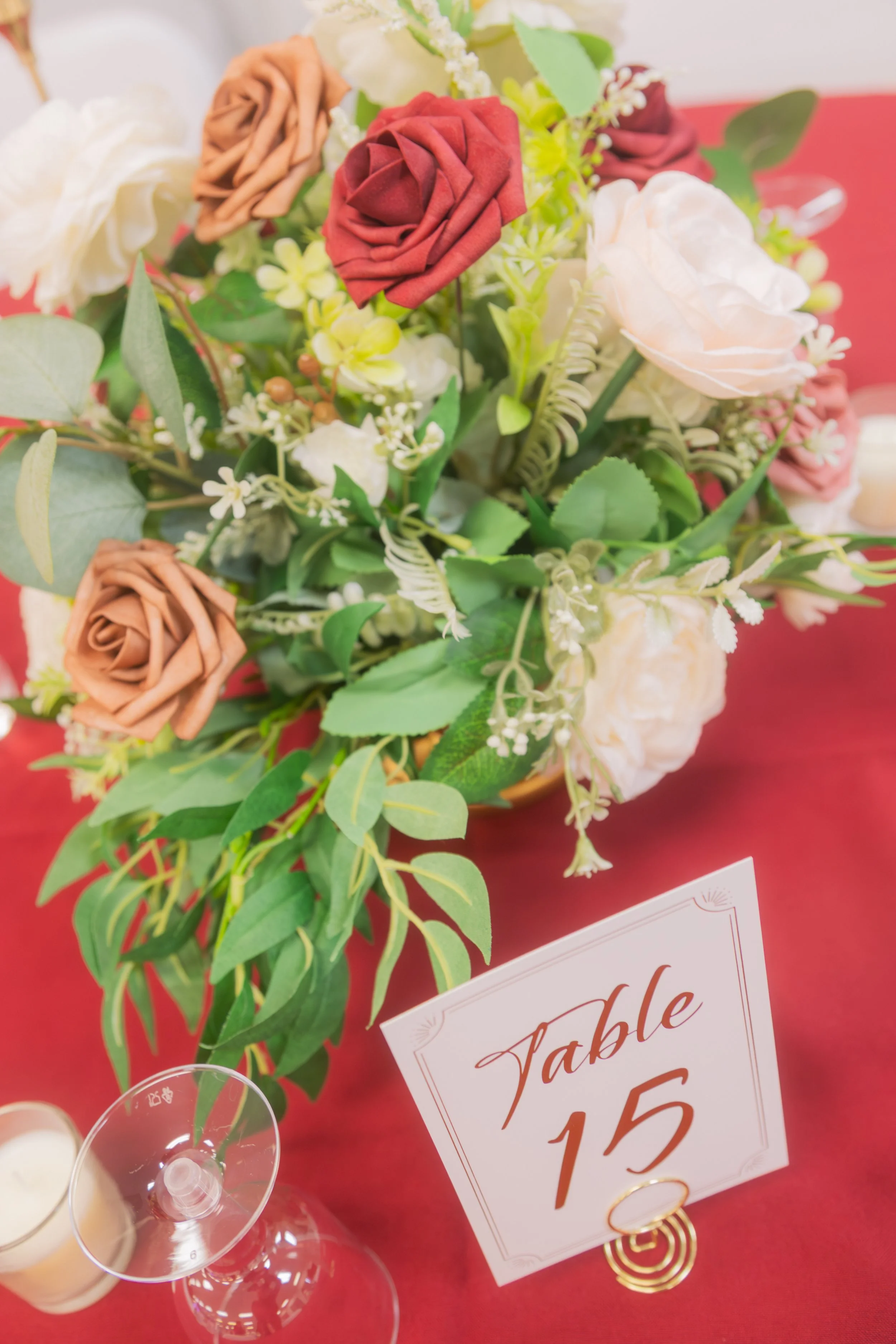 A floral centerpiece with roses and greenery on a red tablecloth, next to a table sign that reads "Table 15."