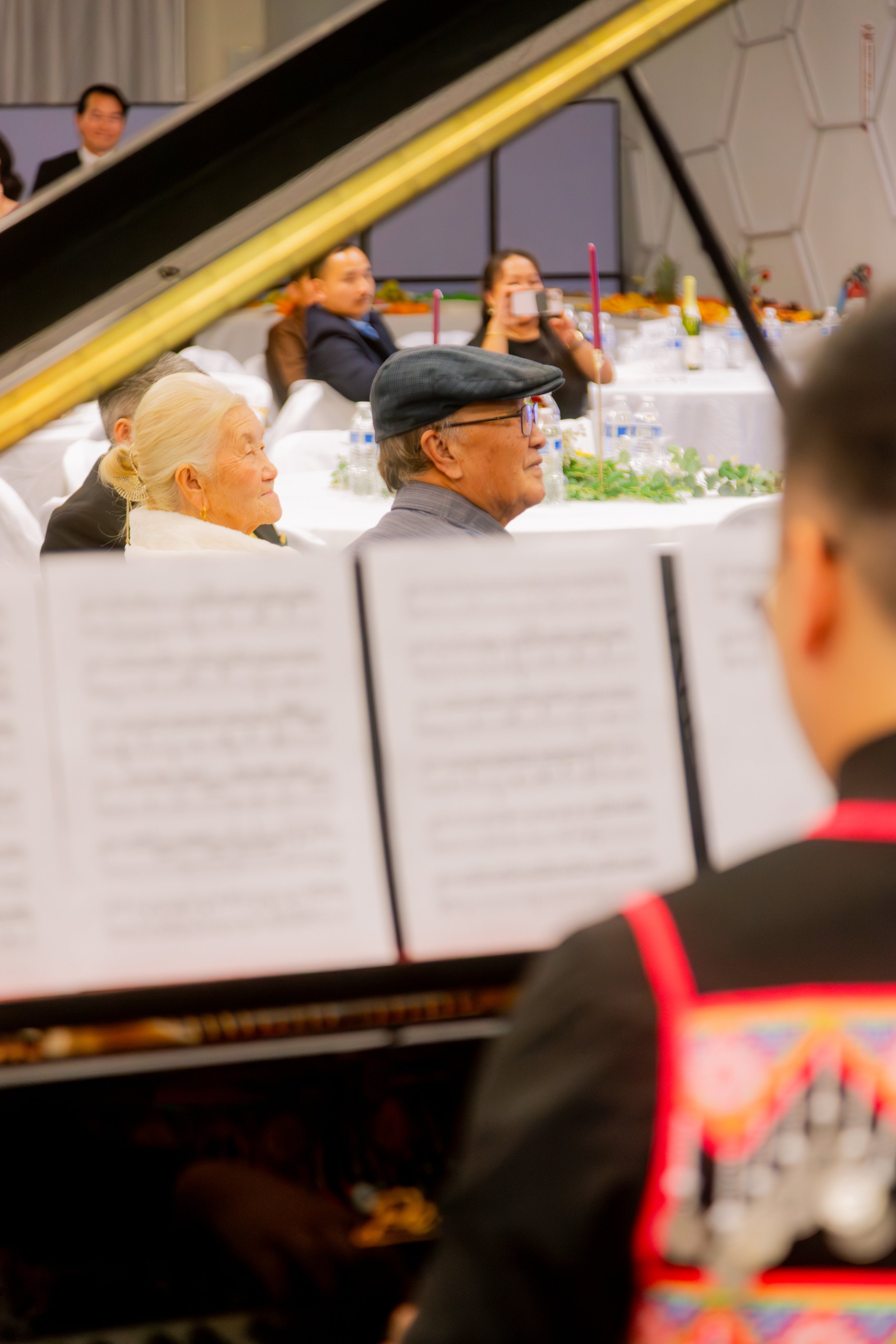 People attending a musical performance, including an elderly woman and a man with glasses and a cap, sitting at tables decorated with candles and greenery, with musicians playing in the foreground and a person taking photos in the background.