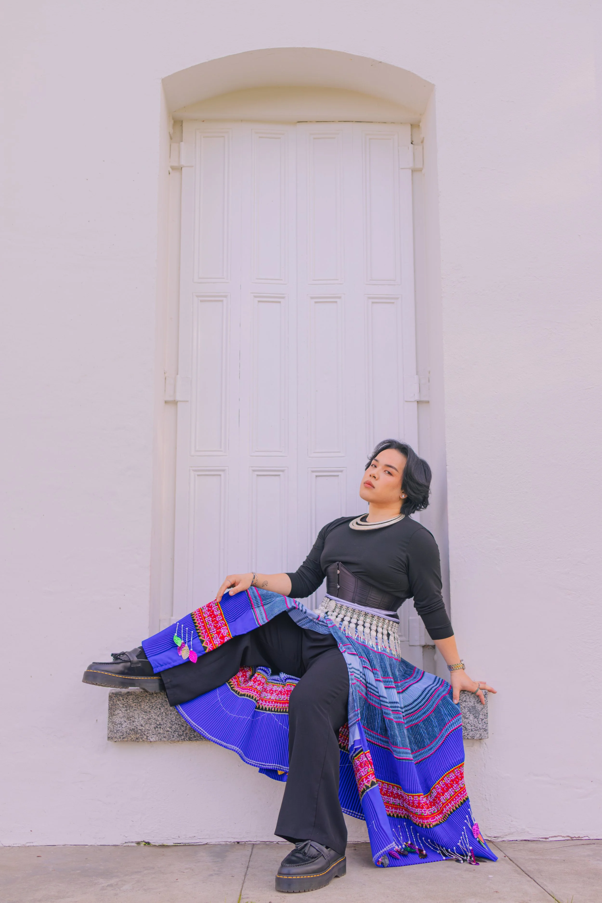 A woman with short black hair sitting on a stone ledge in front of a white wall and closed window with white shutters, wearing a black long-sleeve top, black pants, a wide belt, and a colorful traditional woven skirt with intricate patterns and brigh
