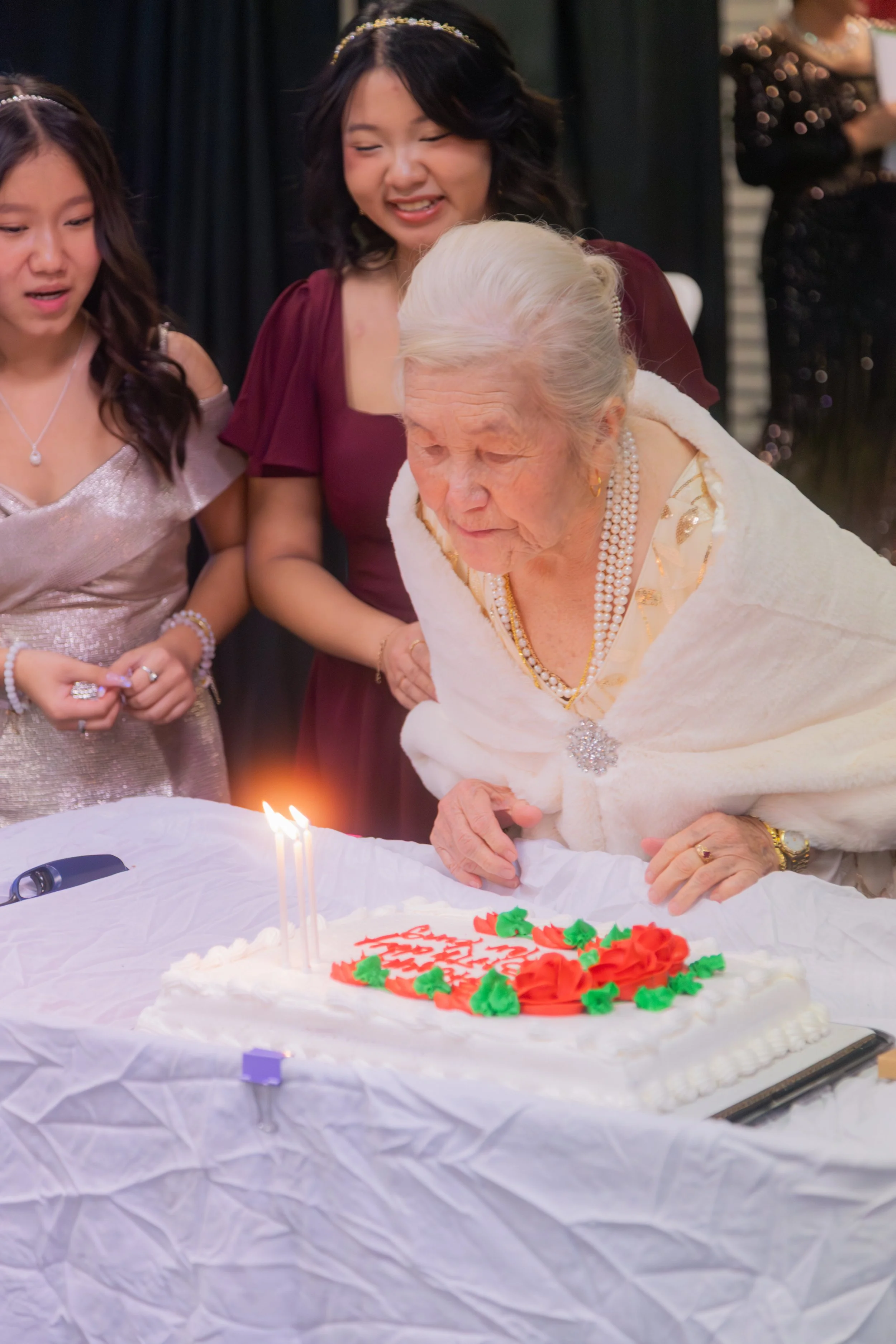 An elderly woman is blowing out candles on a birthday cake while surrounded by three women, celebrating her special day.