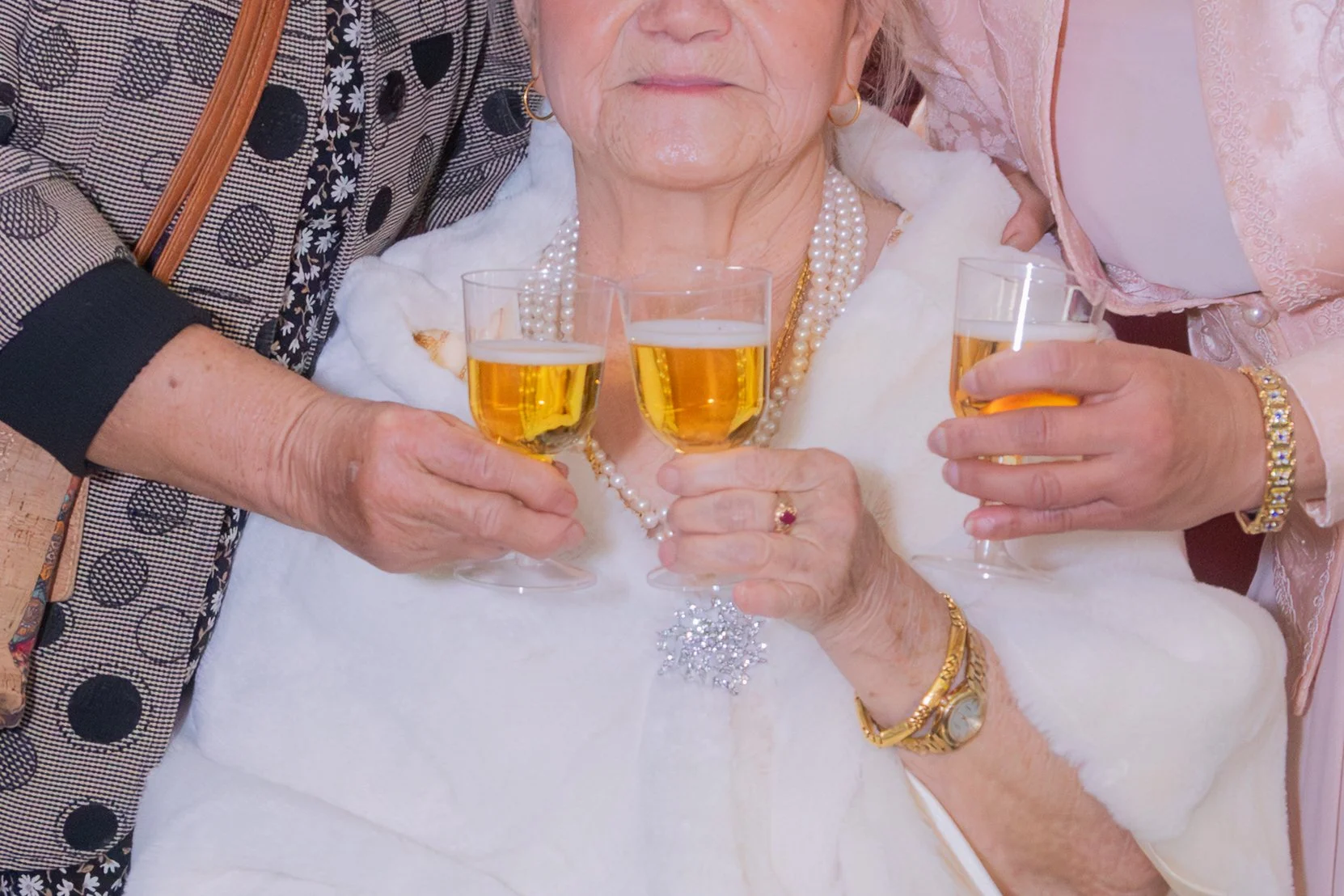 Elderly woman wearing pearl jewelry and a white fur shawl, holding a glass of champagne, surrounded by people celebrating.