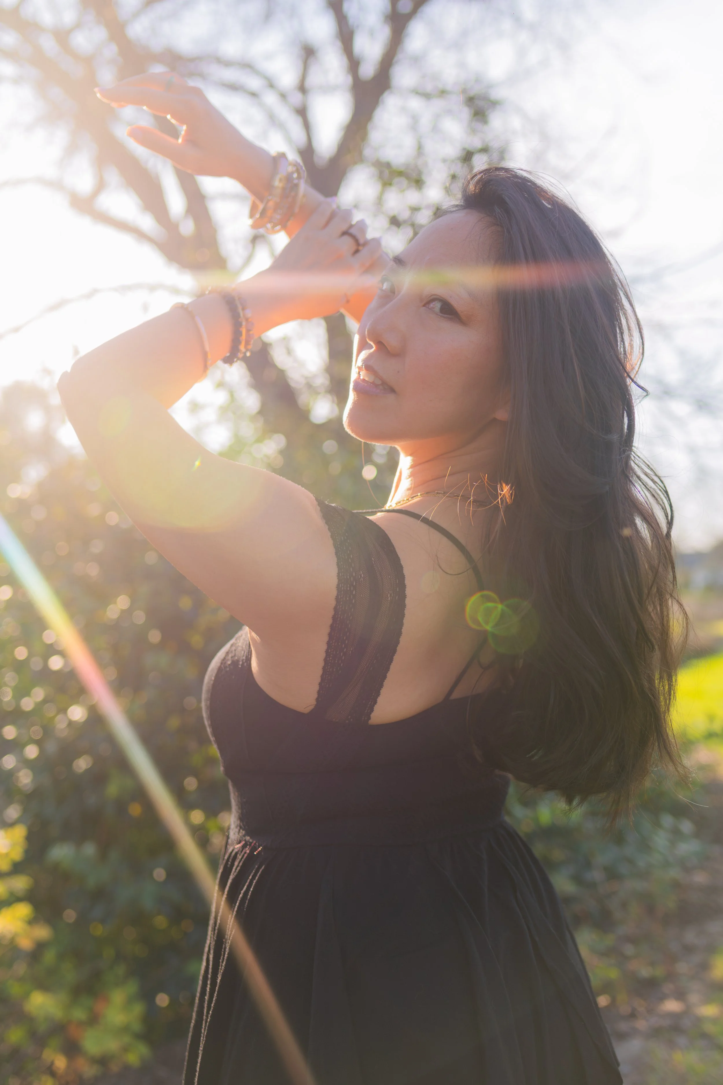 A woman posing outdoors in sunlight, wearing a black dress with lace straps, with trees in the background.