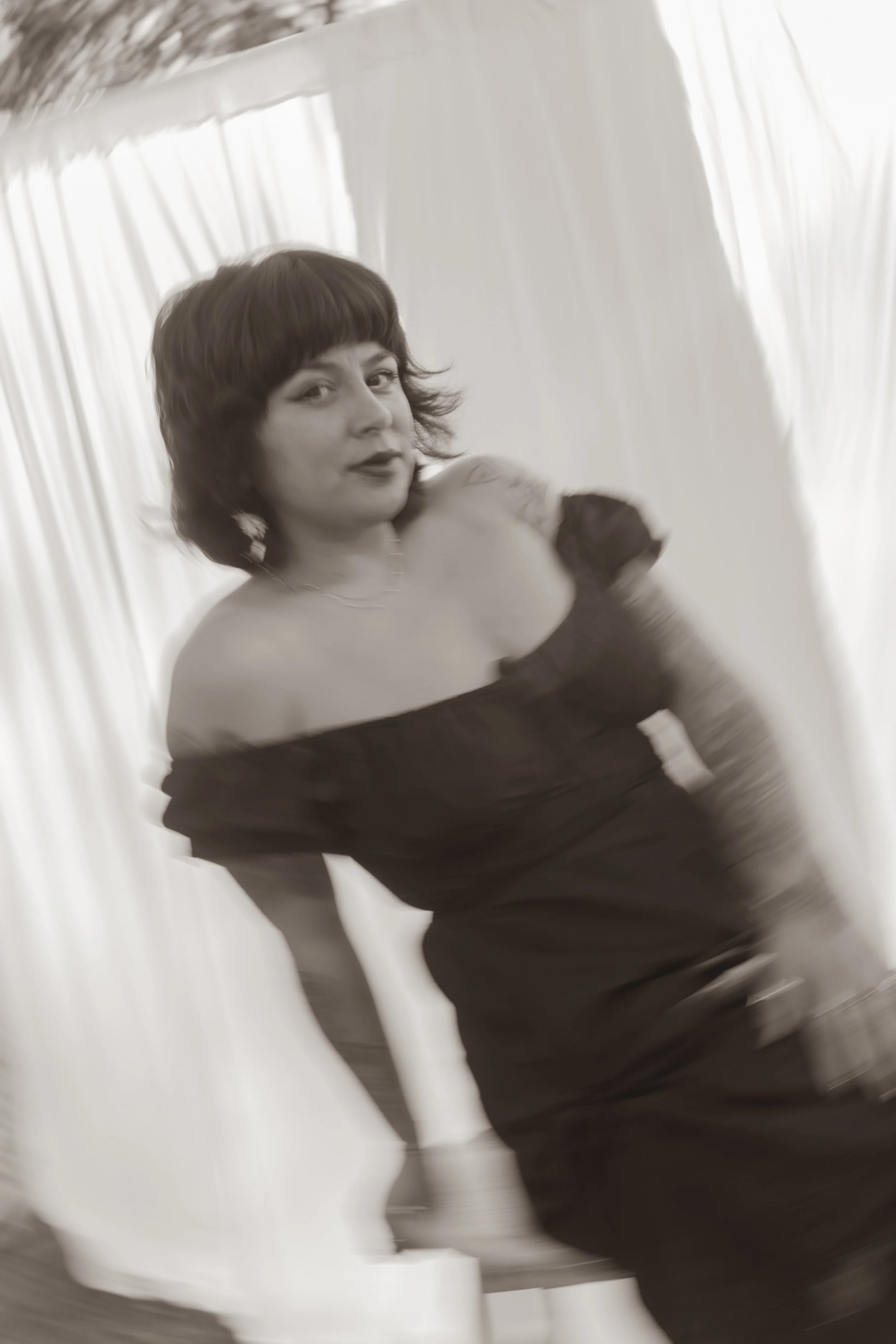 A woman with dark hair in a vintage-style off-the-shoulder dress, sitting on a chair against a backdrop of light-colored curtains.