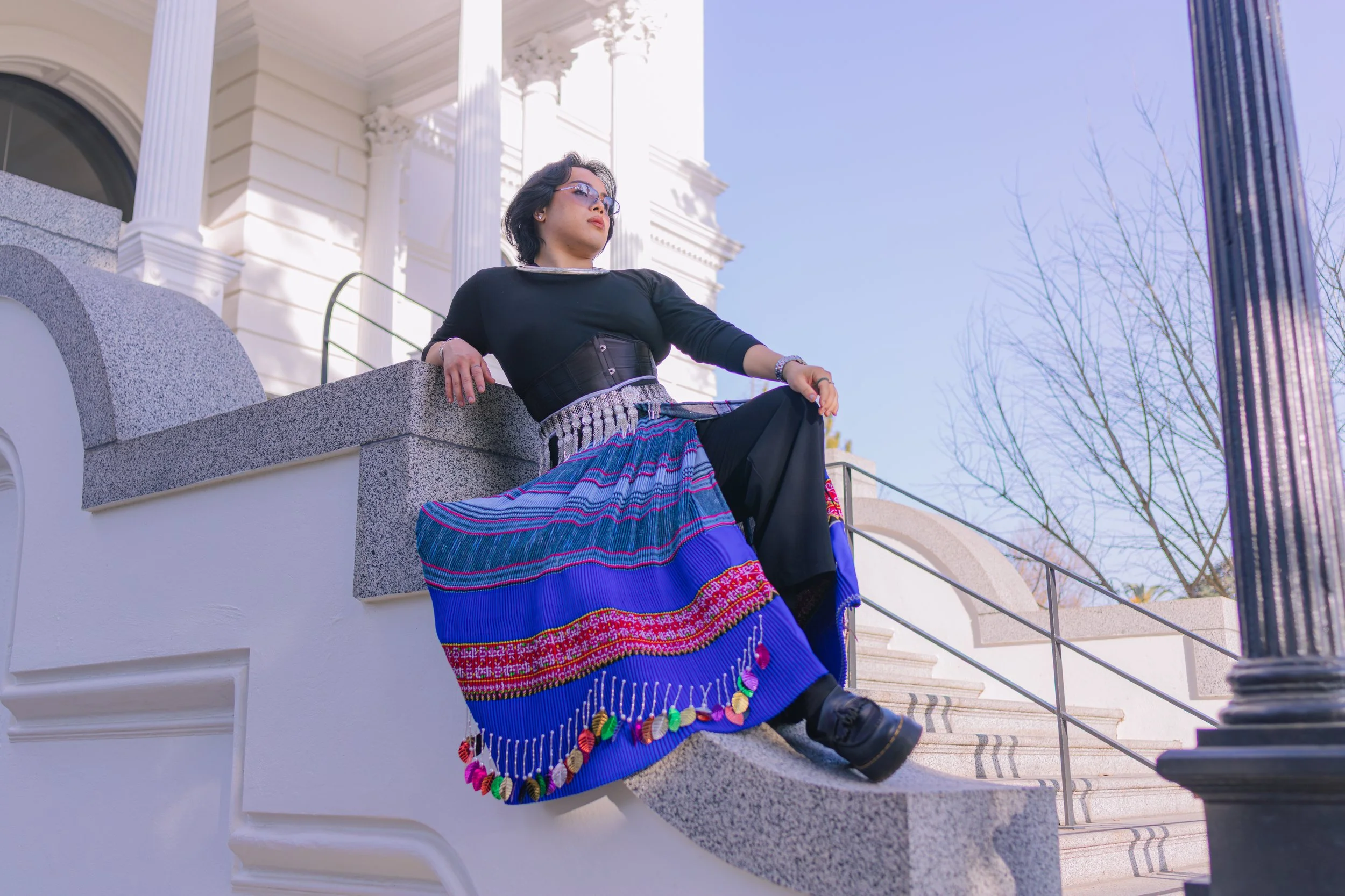 Woman sitting on a stone ledge outside a white building with columns, wearing a colorful skirt, black top, sunglasses, and a watch.