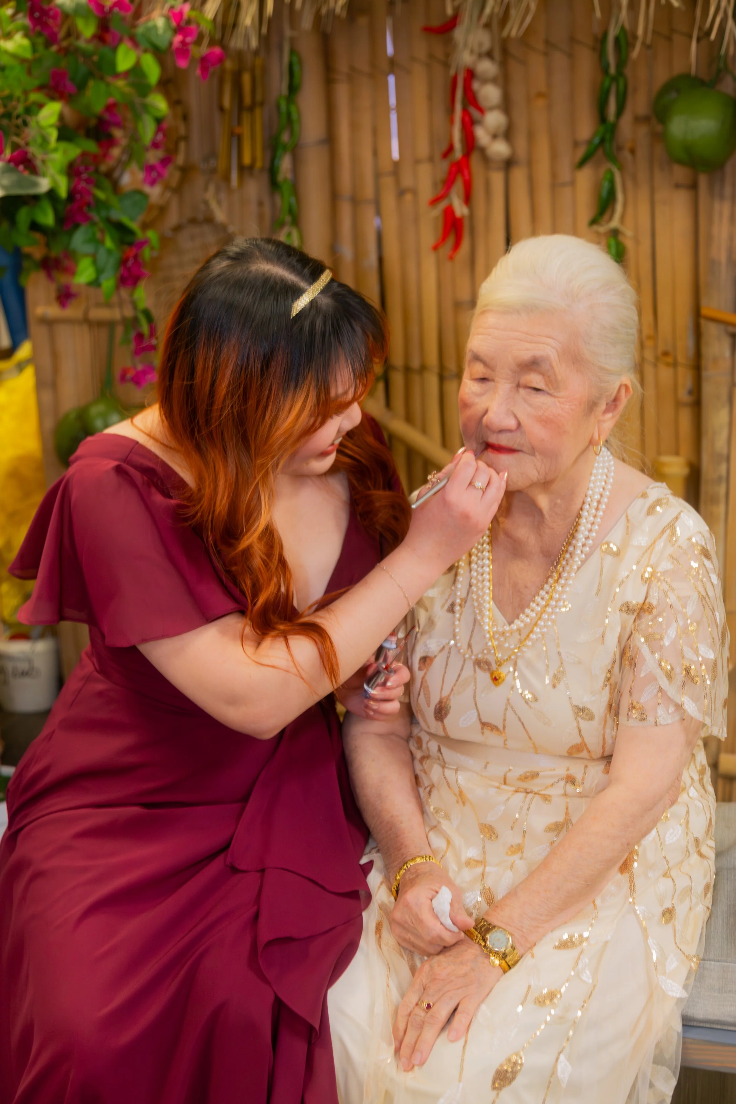 A young woman applying makeup to an elderly woman with white hair, in a decor with bamboo, hanging red chili peppers, and tropical plants.