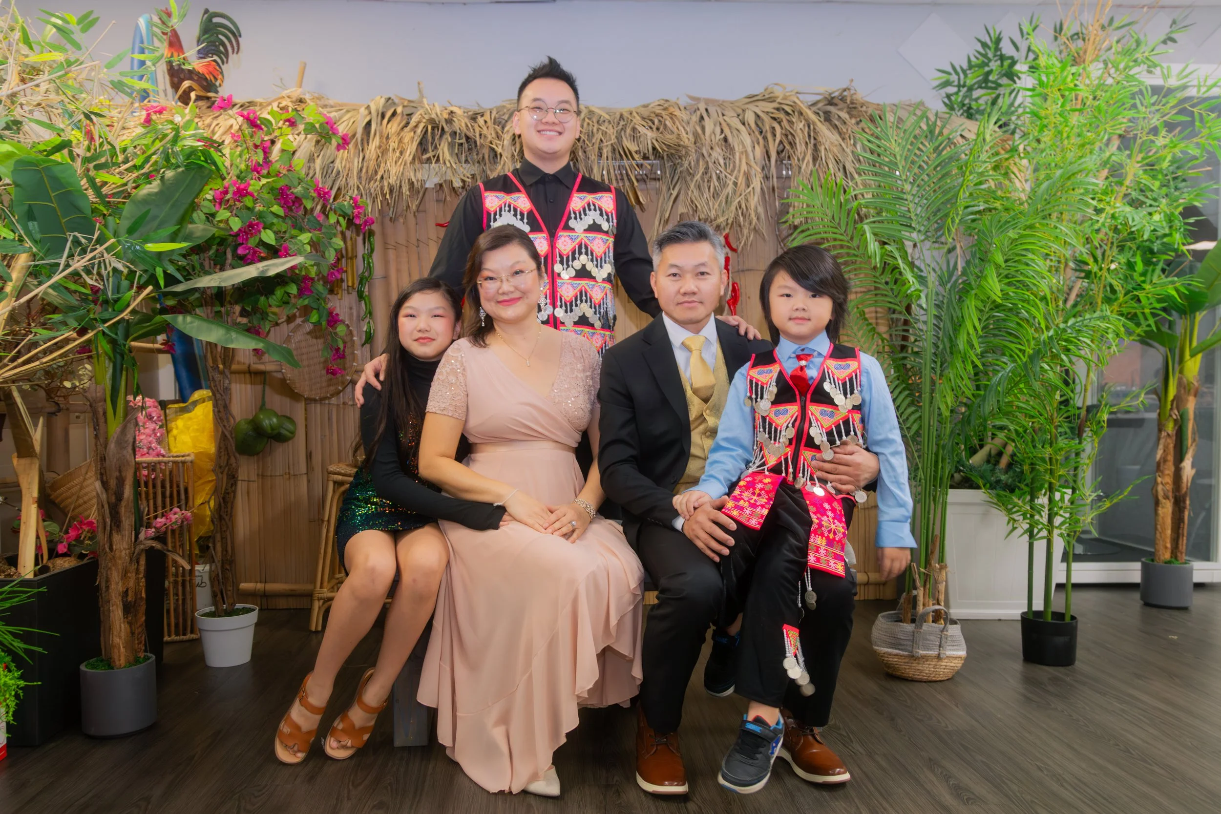 A family of five poses for a photo in front of a tropical-themed backdrop with plants and bamboo decor. Two children and a woman sit in the front, the woman and a boy in traditional attire sitting, while a man and girl stand behind them. The family i