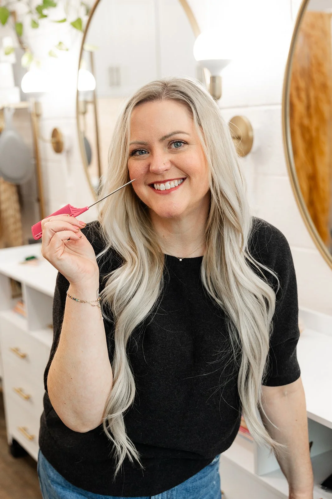 A woman with long, wavy blonde hair smiling while holding a small comb near her face in a well-lit, stylish salon with mirrors and lights.