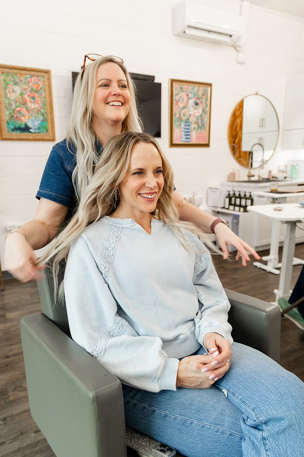 Two women in a hair salon, one sitting in a chair and the other standing behind her, smiling and getting ready to style her hair.