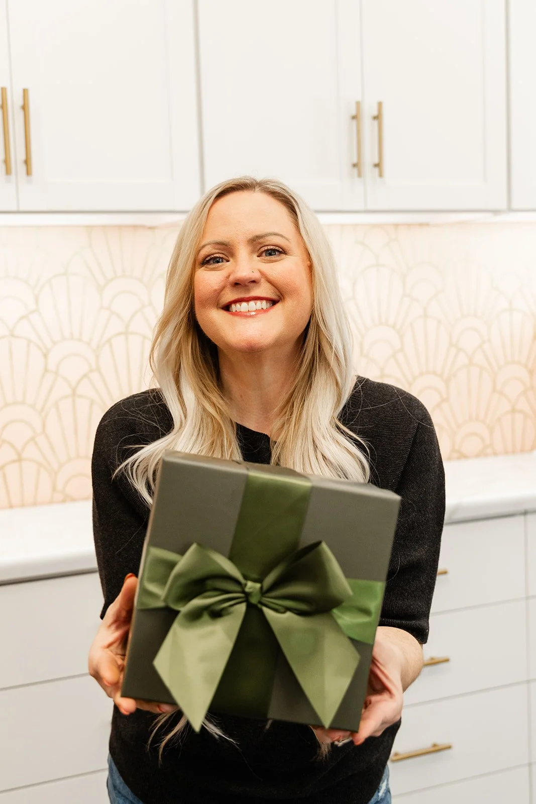 A woman with blonde hair smiling and holding a green gift box with a matching bow, standing in a hair salon
