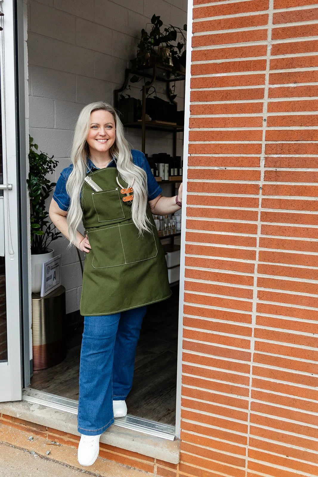 A woman with long blonde hair wearing a blue shirt, green apron, and jeans, standing at the entrance of a salon smiling.