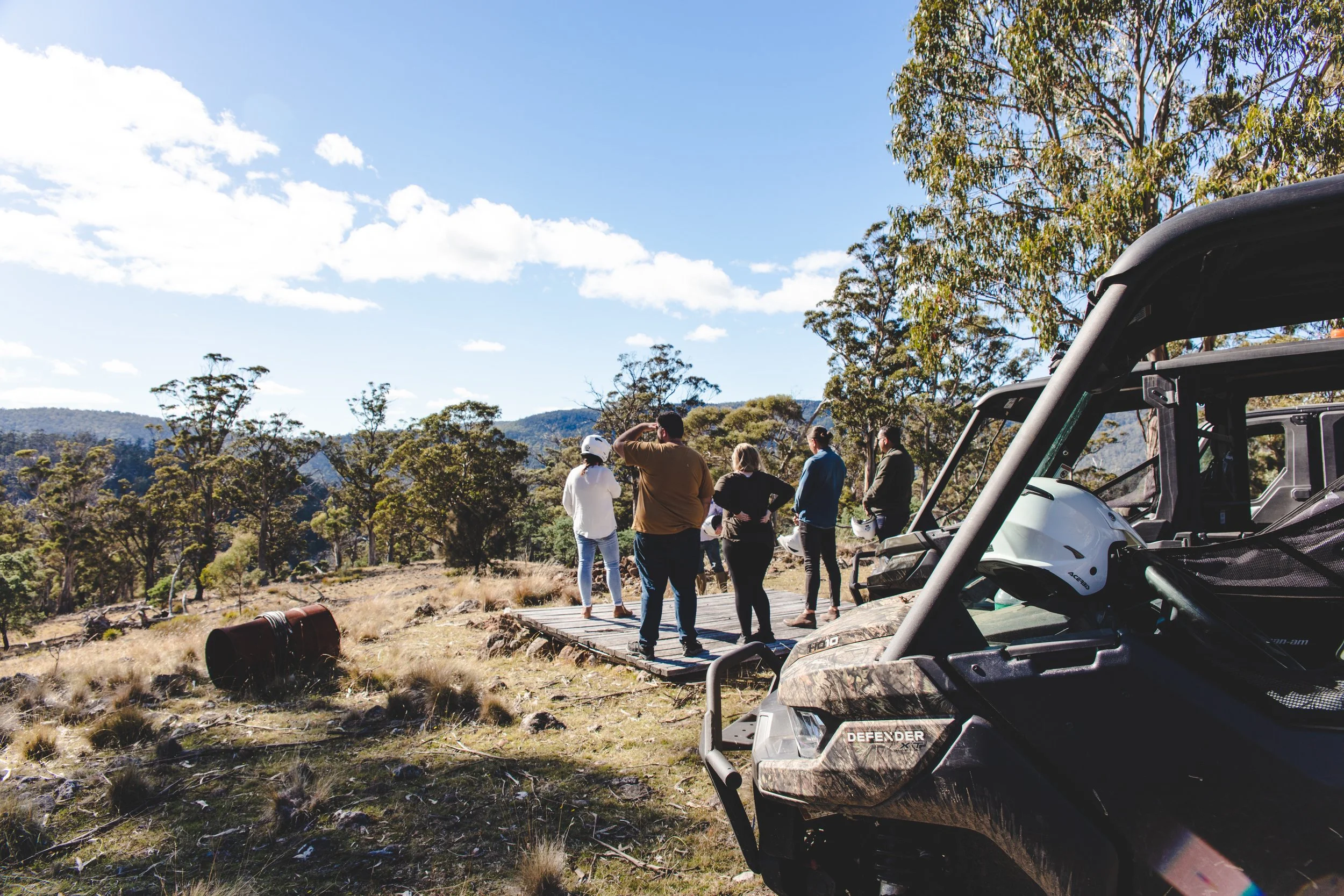 Group of people standing on a wooden platform outdoors, with off-road vehicles and helmets nearby, surrounded by trees and hills under a blue sky with clouds.