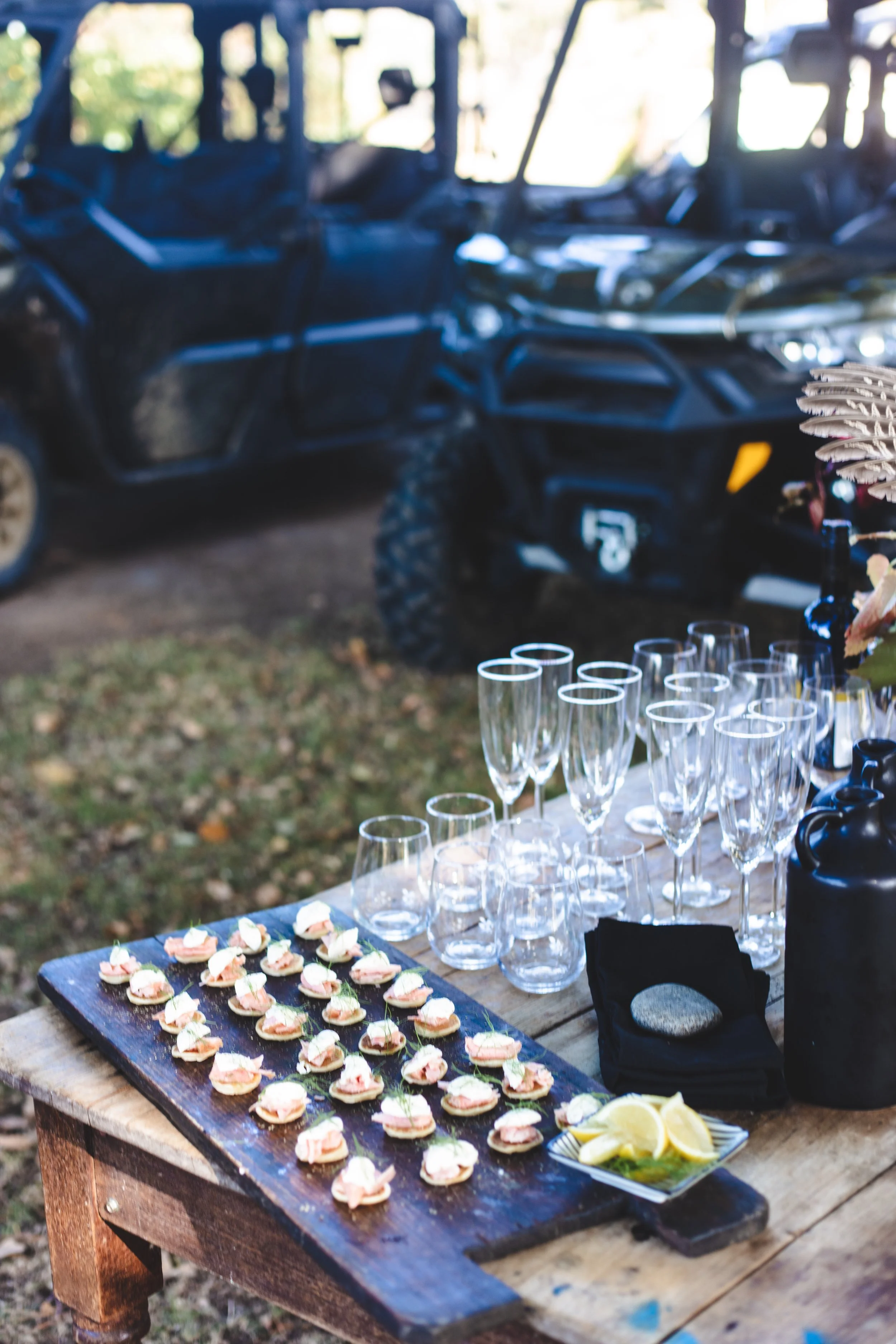 A rustic outdoor table with small appetizers, lemon slices, empty wine glasses, and black napkins, with off-road vehicles in the background.