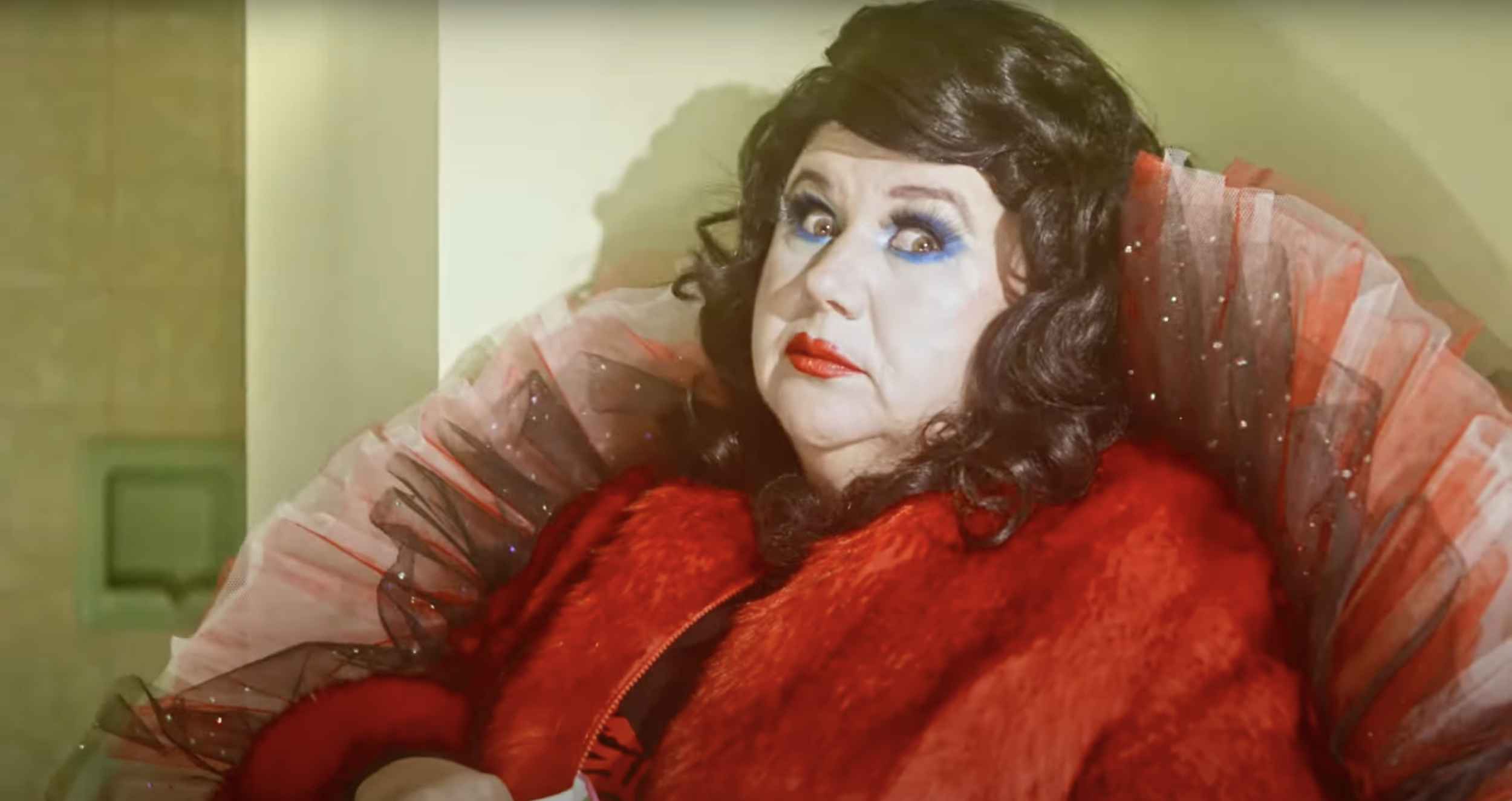A woman with dark curly hair, bold blue eye makeup, and red lipstick, wearing a red fluffy jacket and a large, sparkly, red and black tulle dress, sitting in a chair against a yellow wall.