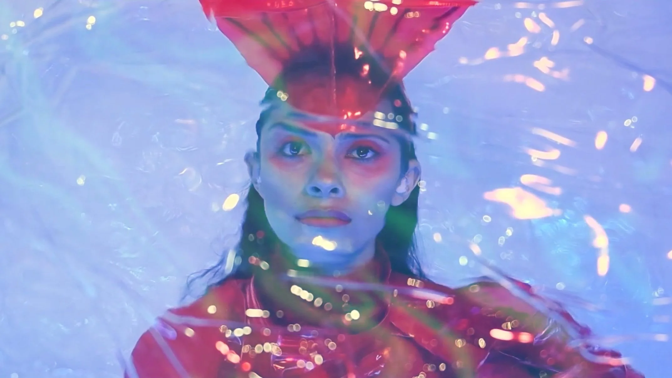 A woman with a serious expression, underwater, with water reflecting colorful lights, wearing a red top and a shiny headband.
