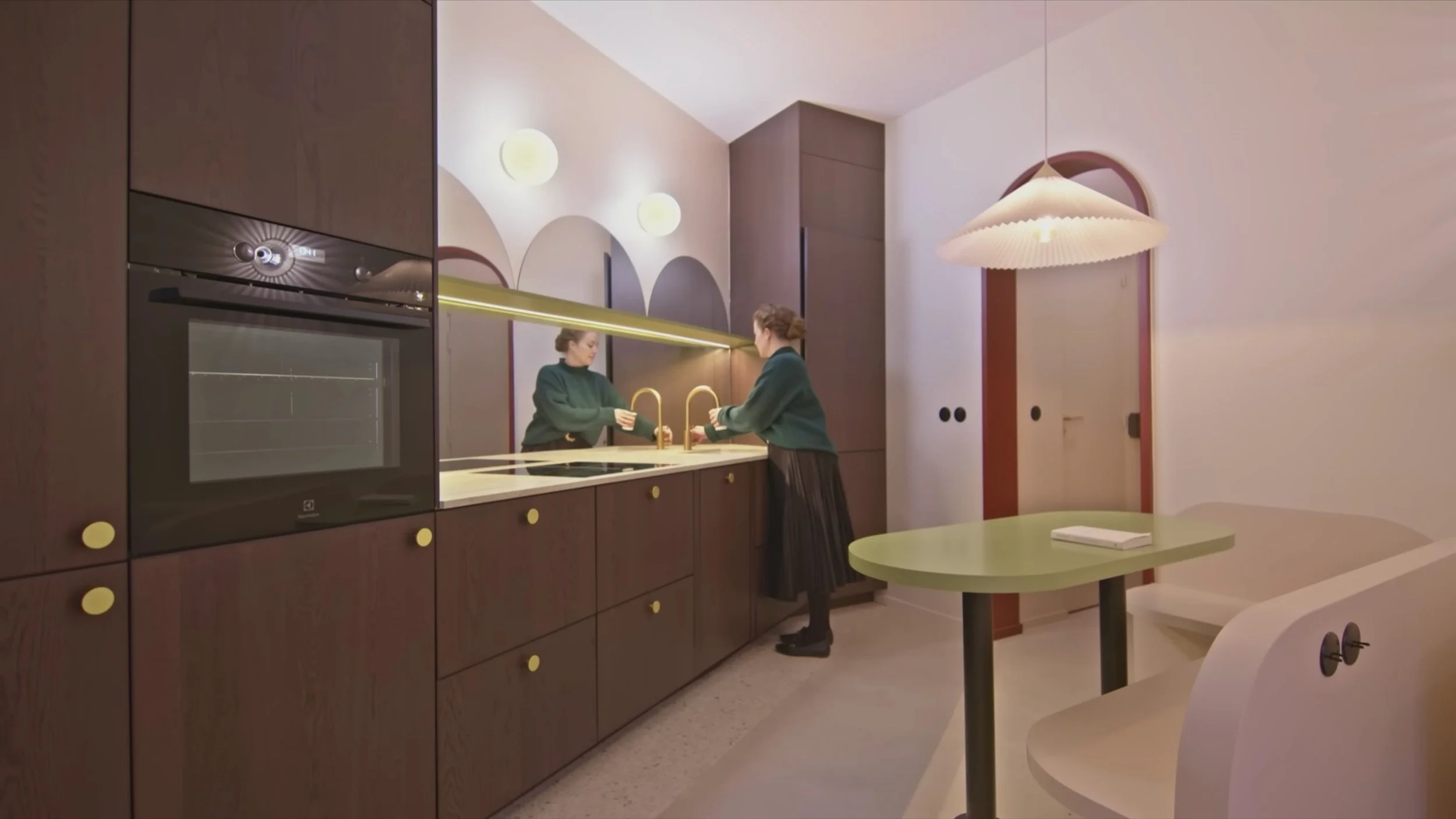 A woman in a green sweater and black skirt stands at a modern kitchen sink, looking in a mirror. The kitchen has dark brown cabinets with round yellow handles, a built-in oven, and a yellow countertop. A round, green table with a white book and a white curved chair are in the foreground. There are arched wall accents, round wall lights, and a hanging ceiling lamp in the background.