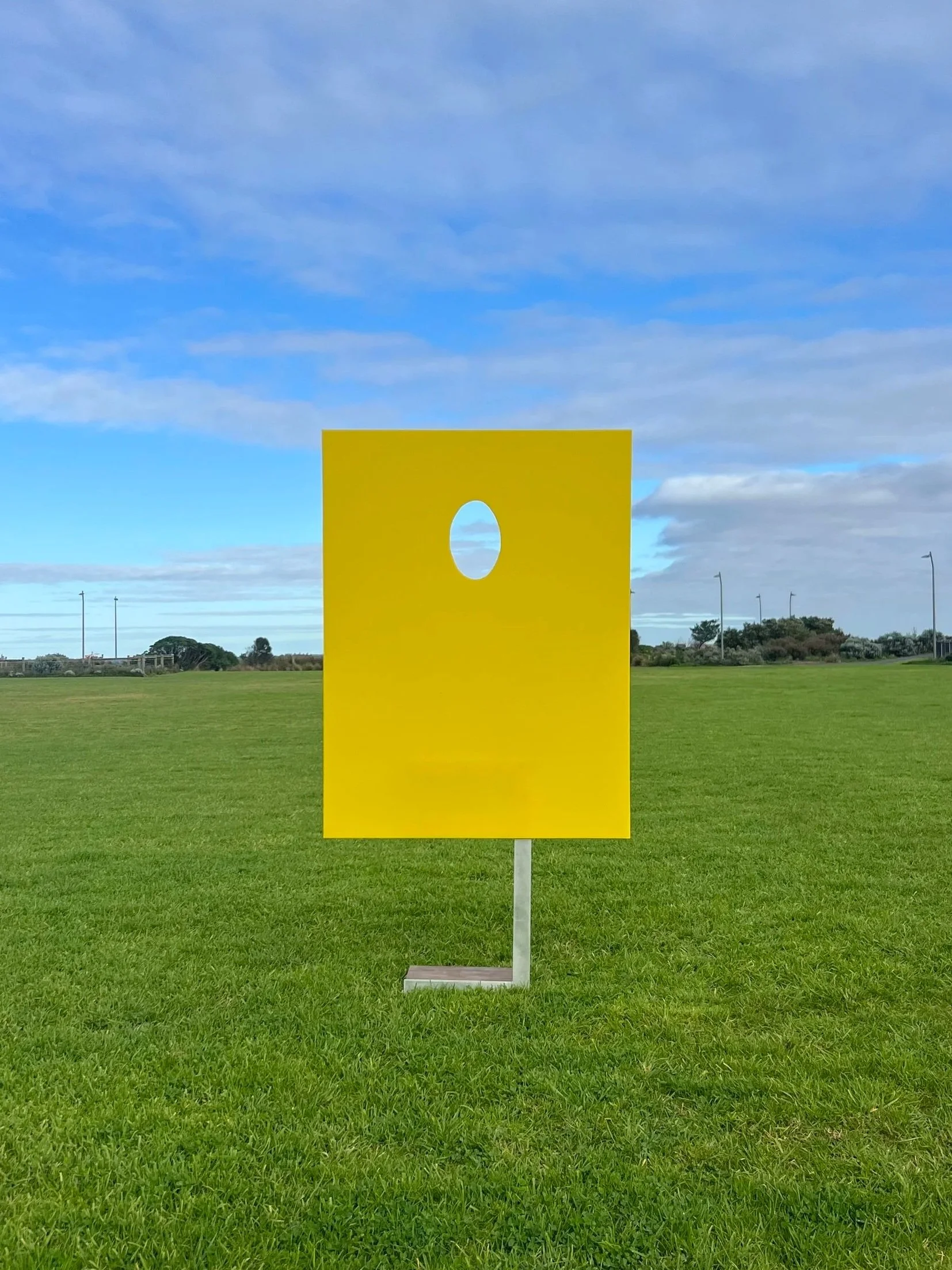 A yellow face-in-a-hole board sculpture, standing on a grassy field with a clear blue sky overhead.