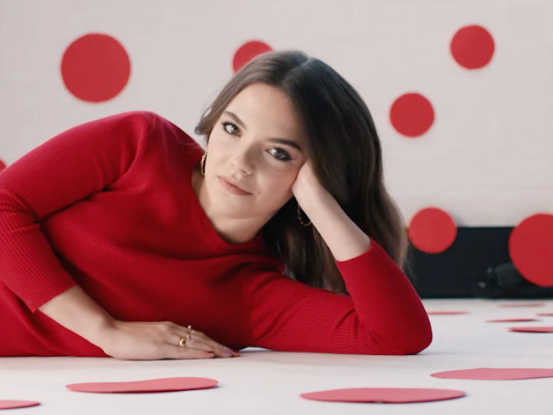 A woman with shoulder-length dark hair wearing a red long-sleeve top, lying on her side with her head resting on her hand, surrounded by red paper circles on a white background.