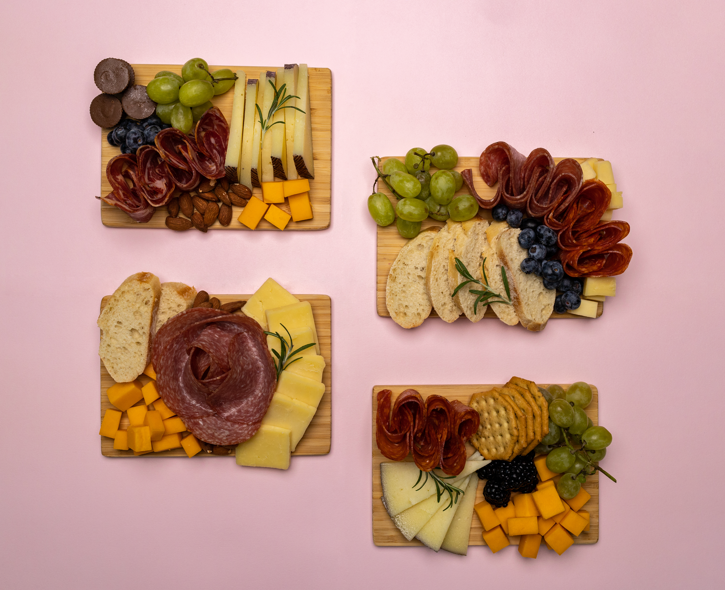 Assorted cheese, sausage, grapes, crackers, blueberries, blackberries, almonds, and some sprigs of rosemary on small wooden boards set on a pink background.