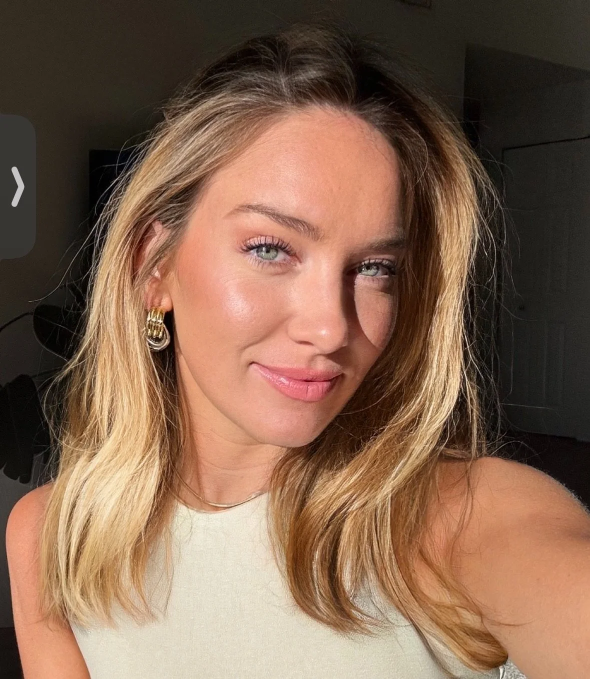 Close-up of a woman with blonde hair and green eyes taking a selfie indoors, wearing a white top and gold hoop earrings, with sunlight illuminating her face.
