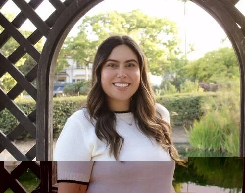Katelyn Askew smiling in a white knit top under a garden archway