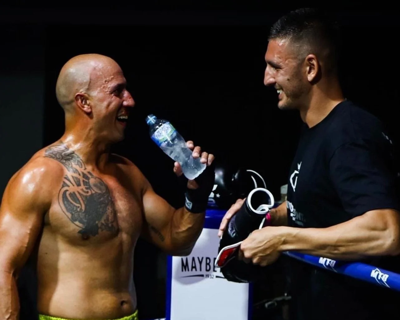 A shirtless male athlete with tattoos on his chest and arm, smiling and holding a water bottle, stands next to a man in black shirt holding gym equipment and a phone, both smiling.