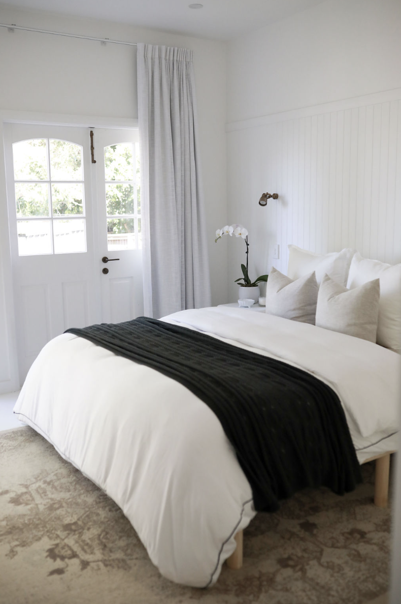 Bright bedroom with white bed, black blanket, and beige pillows. White walls, a window with white curtains, and a potted orchid plant beside the bed.