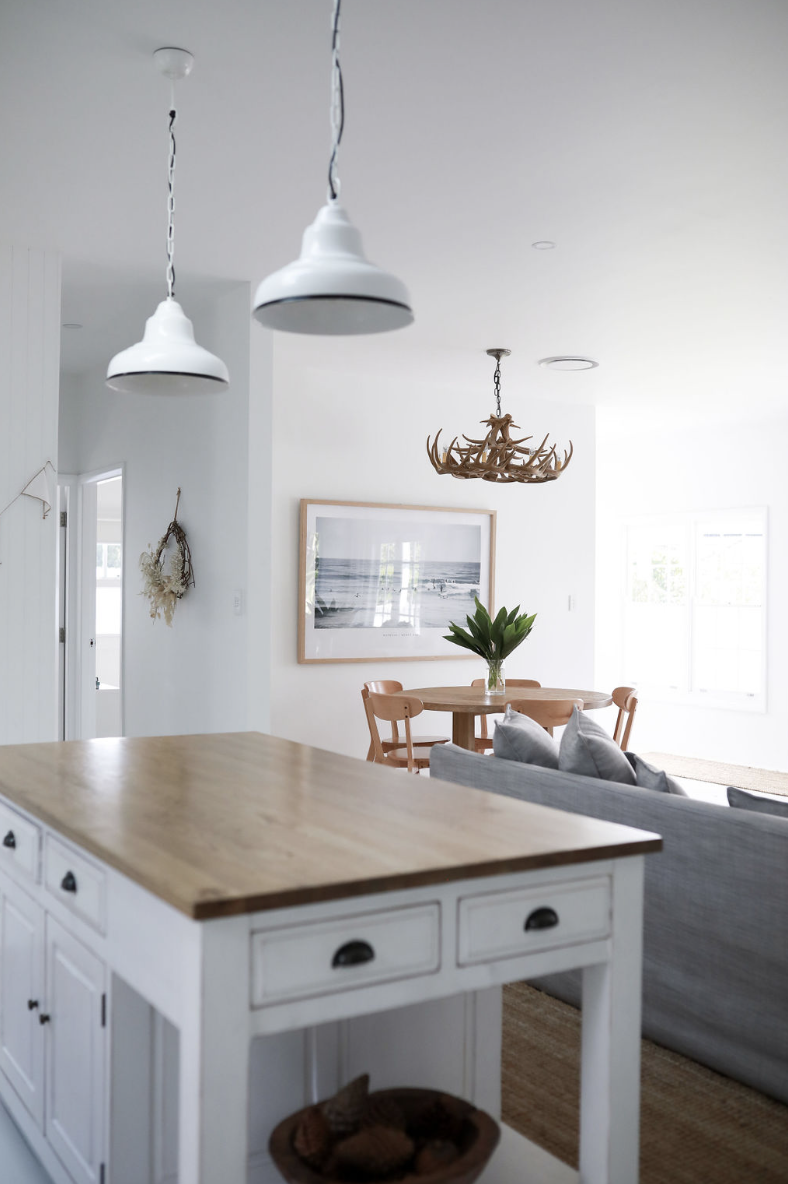 Open-concept living and dining area with white walls, a wooden dining table with chairs, a gray sofa, pendant lights, a framed picture of the beach, a chandelier made of antlers, and a kitchen island with a wooden top.