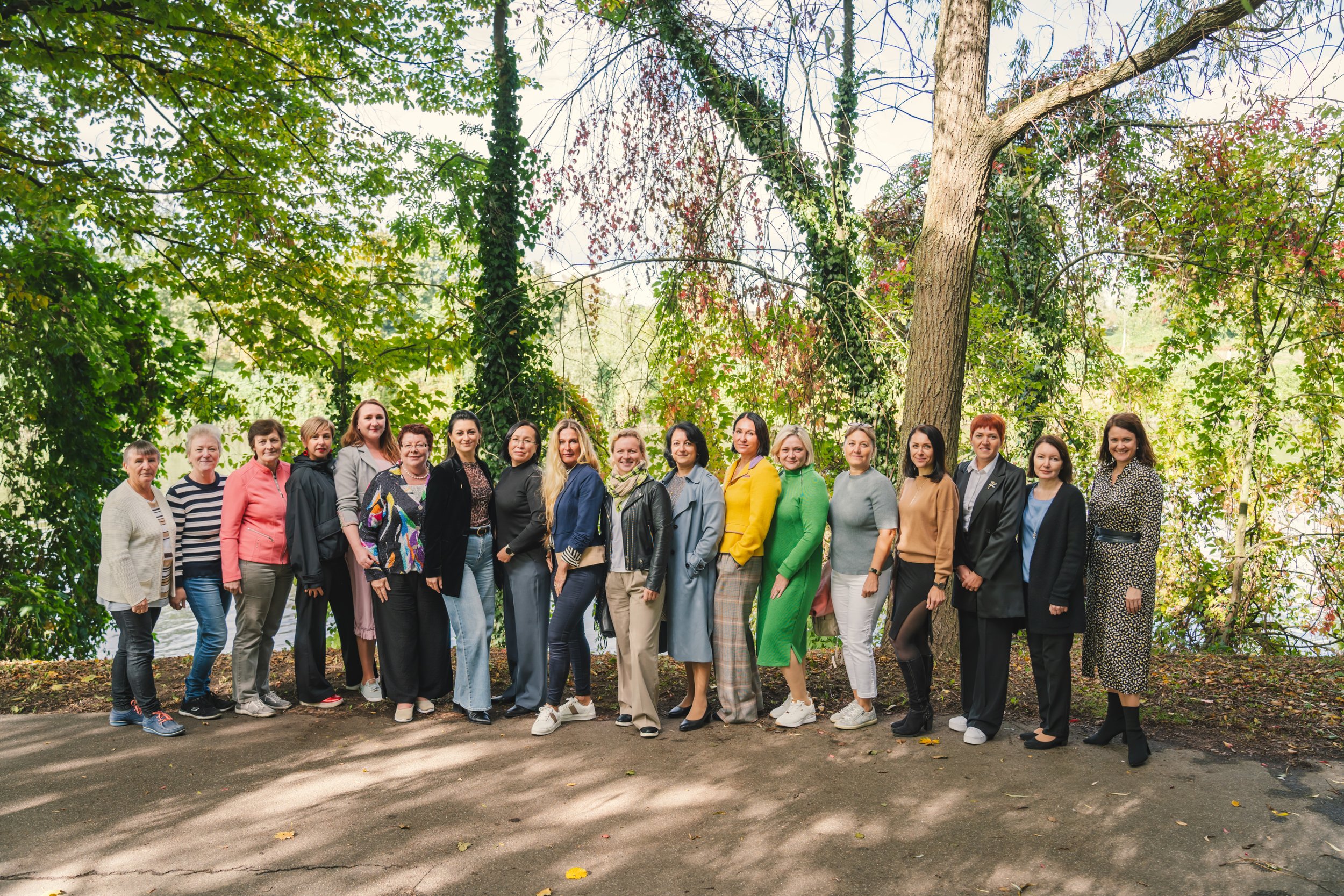 Gruppenfoto von 16 Frauen, stehend im Freien vor einer Baumgruppe, bei Sonnenlicht im Park im Herbst.